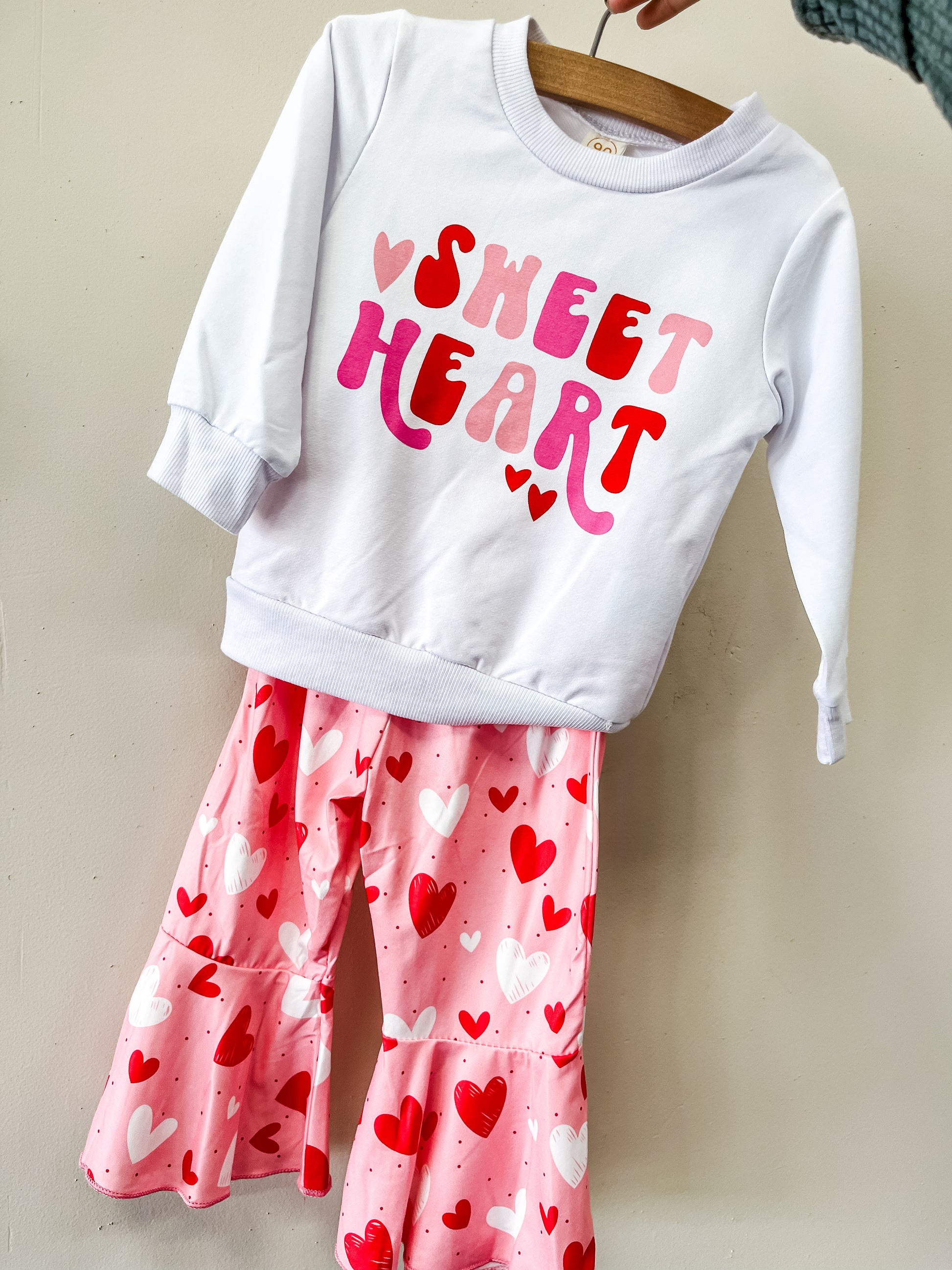 The Sweetheart Vibes Set from Tiny Tot Bundles includes a white long-sleeve top with "Sweet Heart" in pink and red, paired with pink flared pants featuring red, pink, and white heart designs.