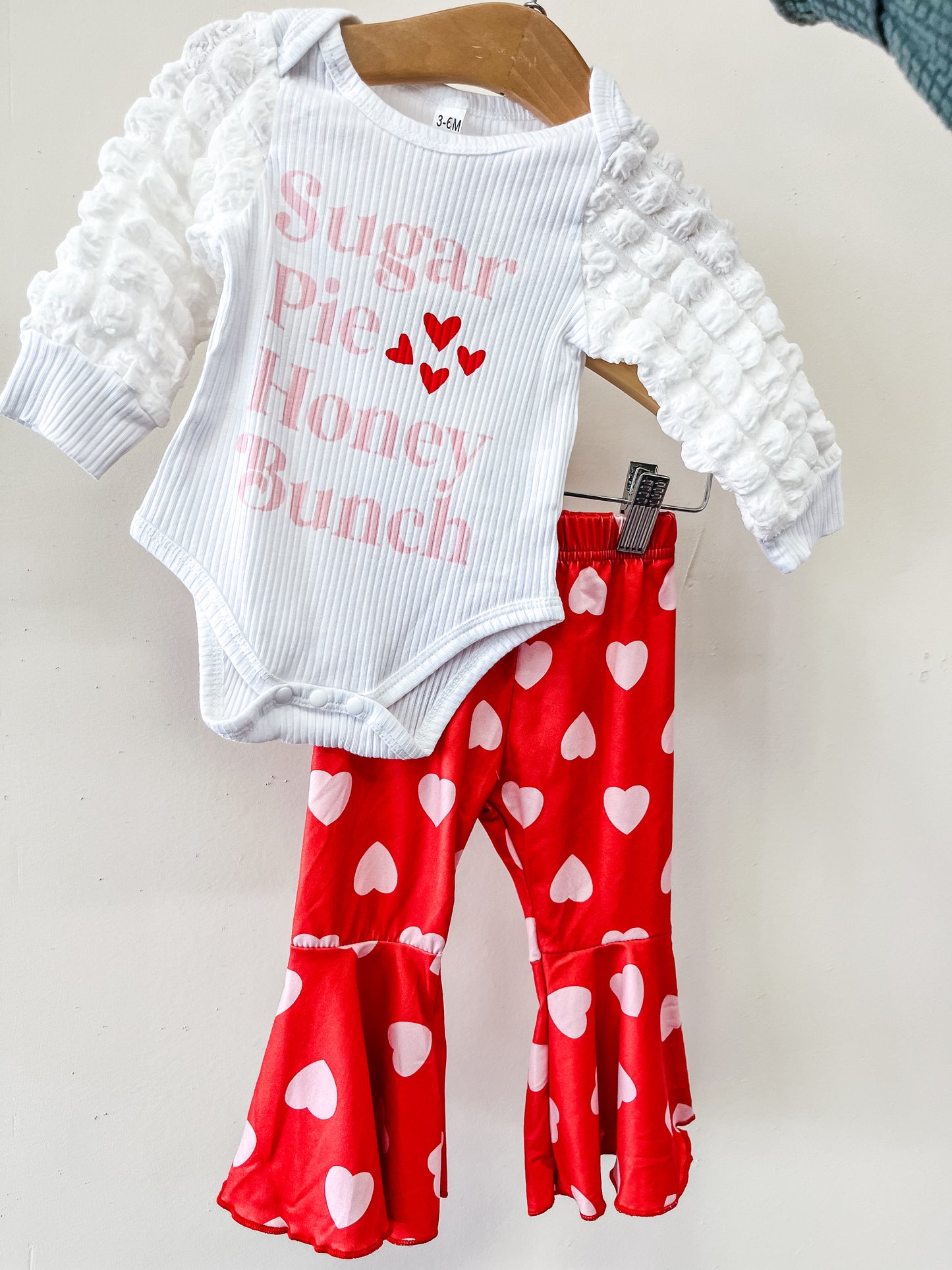 The Sugar Pie Honey Bunch Set by Tiny Tot Bundles includes a white long-sleeve onesie with pink "Sugar Pie Honey Bunch" text and red hearts, plus red bell-bottom pants with white heart patterns, displayed on a hanger.