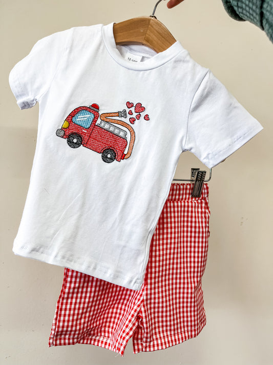 The Heart Hero Firetruck Set by Tiny Tot Bundles features a white child’s T-shirt with an embroidered red fire truck and hearts, paired with red and white checkered shorts, shown on hangers against a plain background.