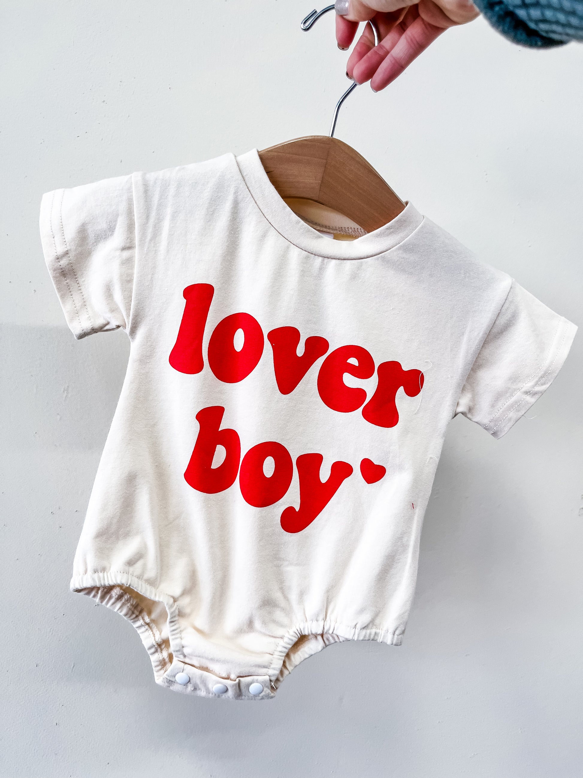 A hand holds the Beige Lover Boy Bubble by Tiny Tot Bundles on a wooden hanger. Bold red text on the front reads "lover boy" with a small heart beside it.