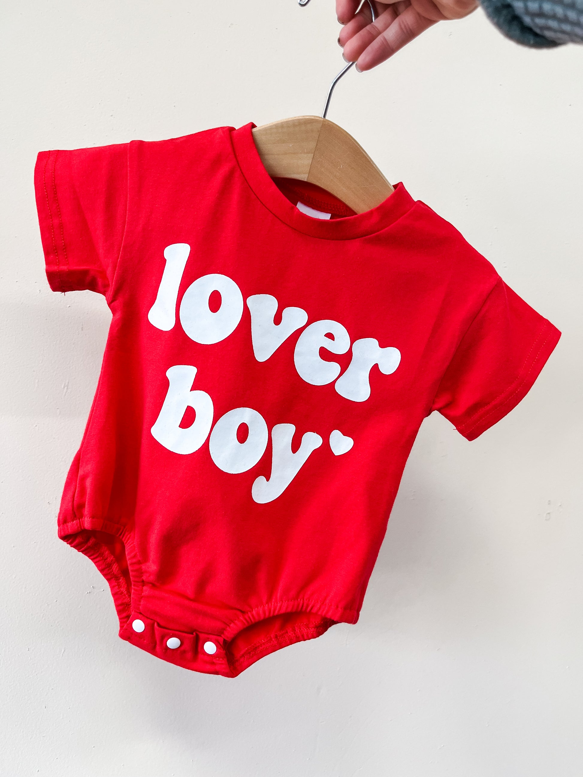 The Red Lover Boy Bubble by Tiny Tot Bundles is a short-sleeve red baby onesie with white snaps and bold “lover boy” text with a heart on the front, shown on a wooden hanger.