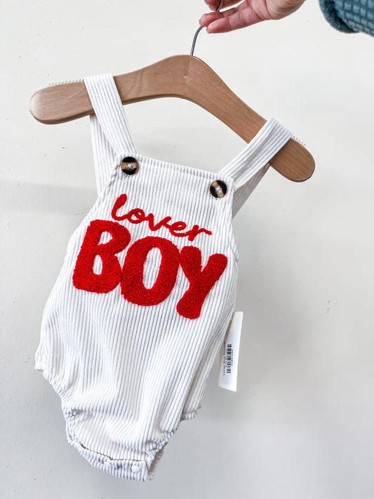 The Lover Boy Romper by Tiny Tot Bundles, a white baby romper with thick straps and two buttons, hangs on a wooden hanger. Bold red “lover BOY” lettering is on the front as someone holds it against a plain white background.