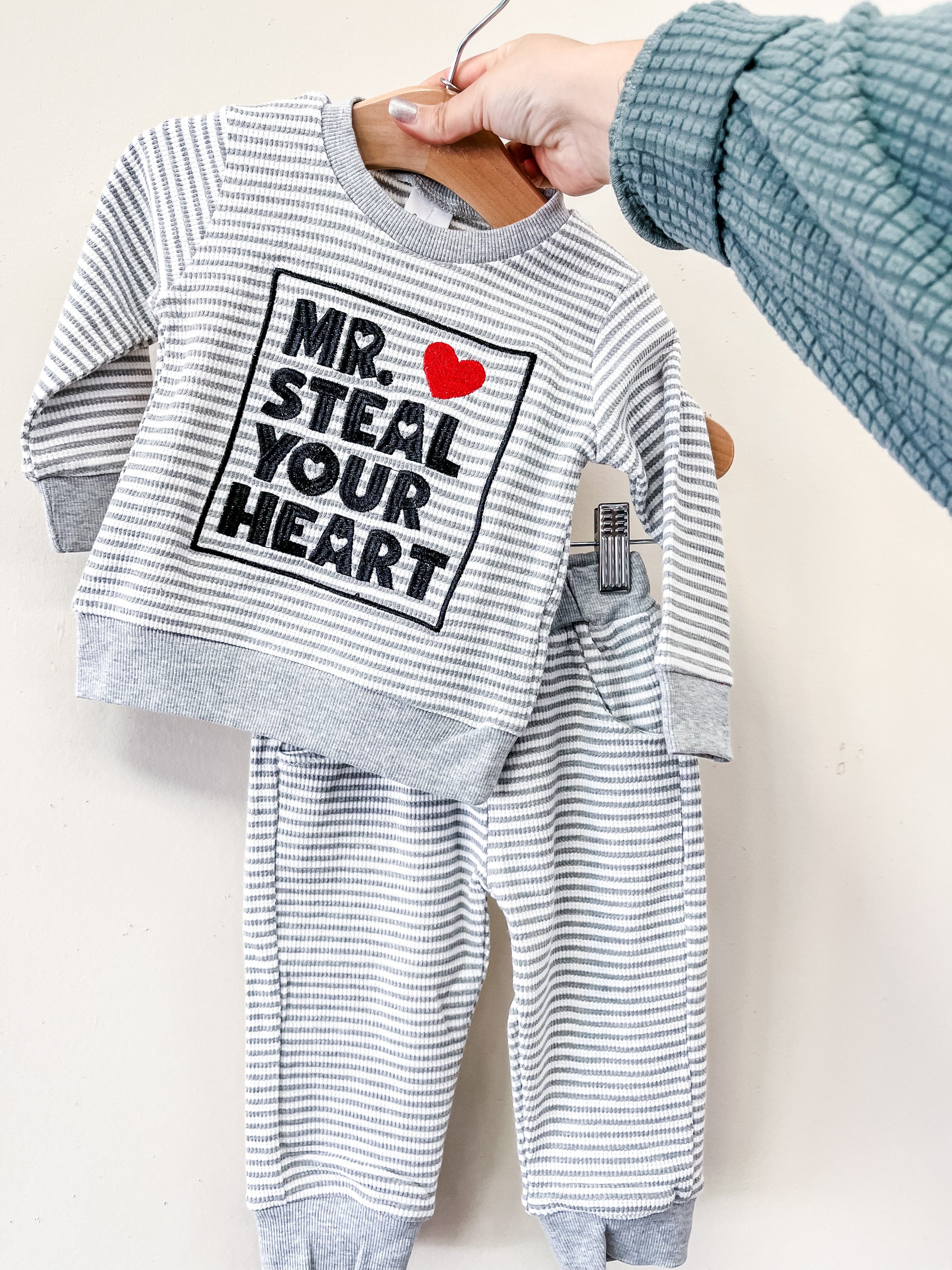 A hand displays the Little Hearts Stealer Set by Tiny Tot Bundles—a gray and white striped baby outfit with a long-sleeve top featuring "MR. STEAL YOUR HEART" and a red heart, plus matching pants.
