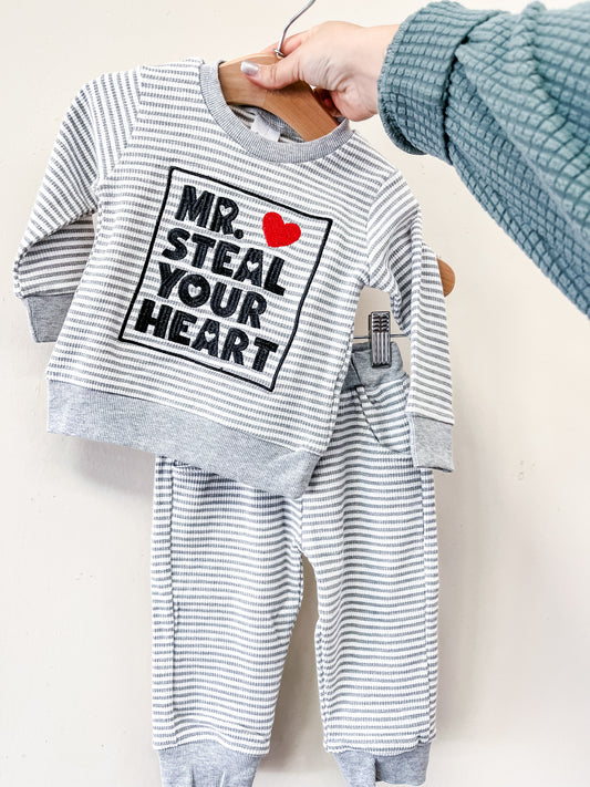 A hand displays the Little Hearts Stealer Set by Tiny Tot Bundles—a gray and white striped baby outfit with a long-sleeve top featuring "MR. STEAL YOUR HEART" and a red heart, plus matching pants.
