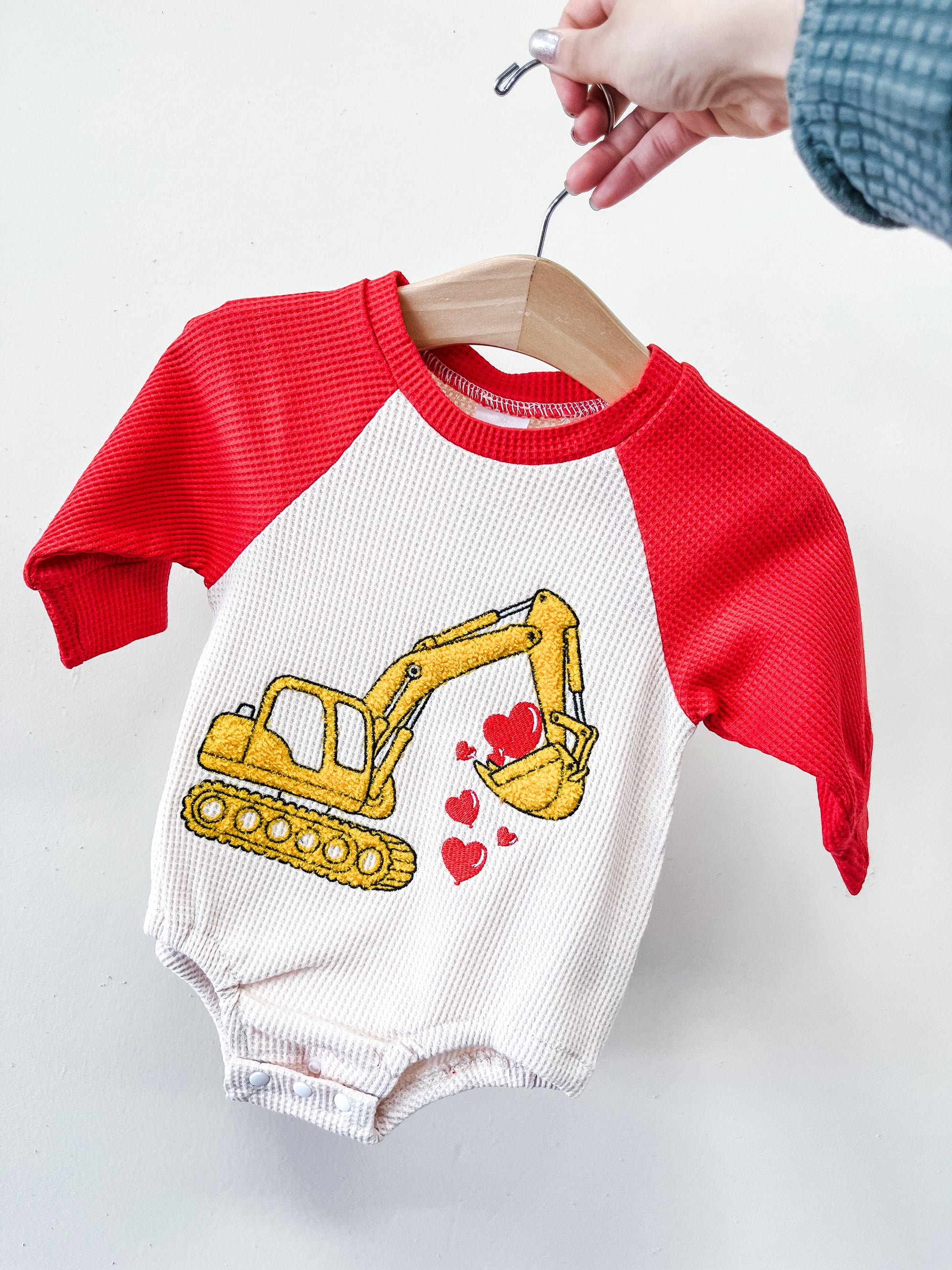 A hand holds a hanger with Tiny Tot Bundles' Digger Of My Heart Romper, featuring red sleeves and a white body with a yellow excavator scooping red hearts.
