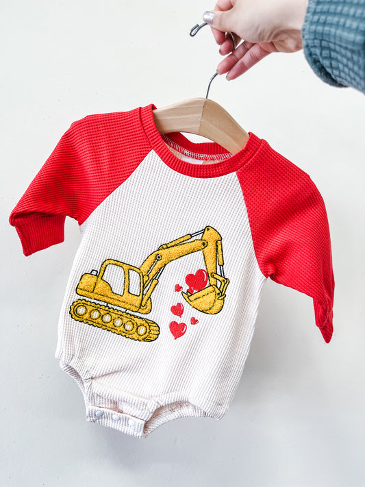 A hand holds a hanger with Tiny Tot Bundles' Digger Of My Heart Romper, featuring red sleeves and a white body with a yellow excavator scooping red hearts.