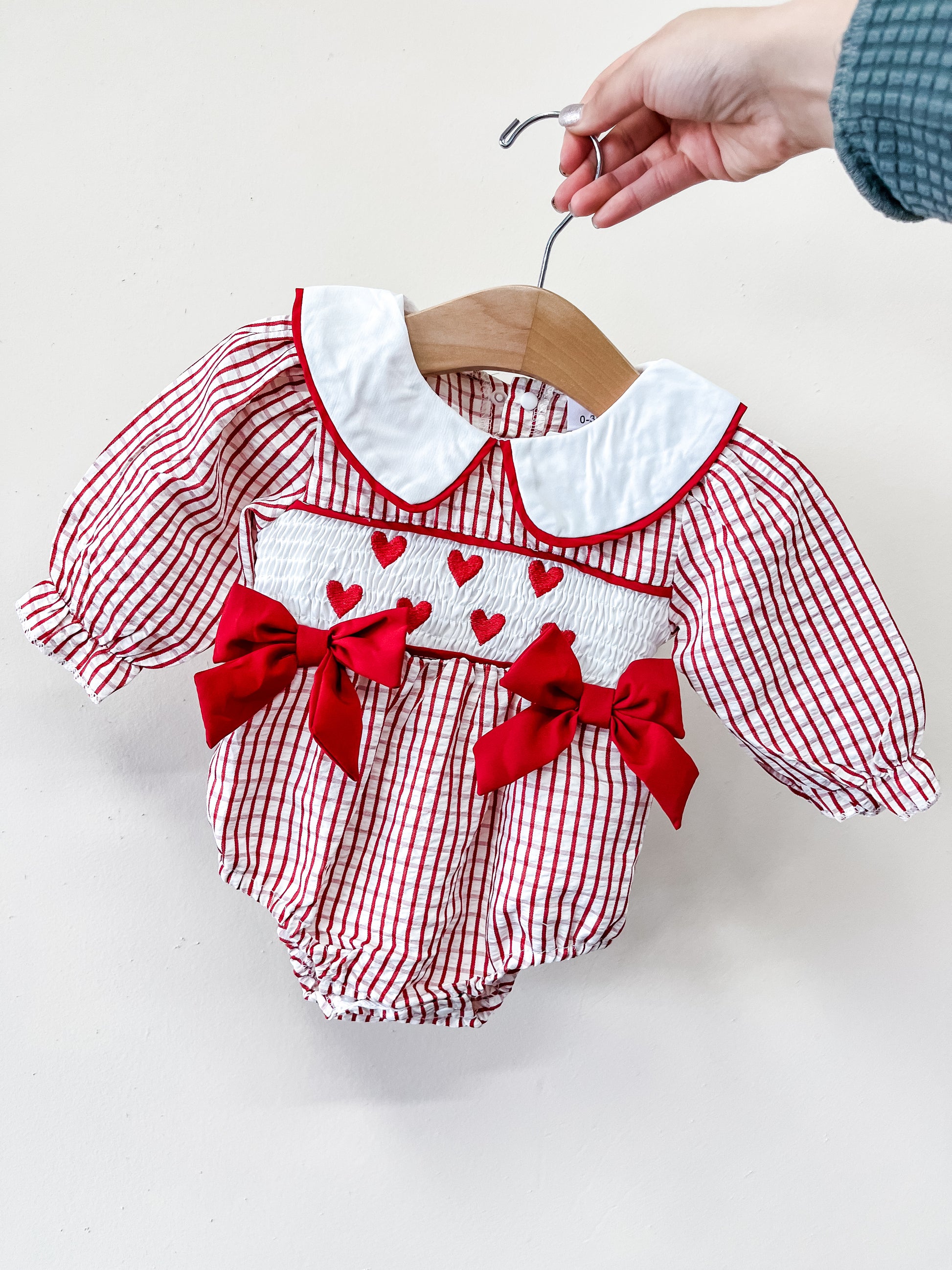 A hand holds up Tiny Tot Bundles' Sweetheart Gingham Bow Romper, featuring white fabric with red vertical stripes, a white collar, red bows, and a row of red hearts across the chest.