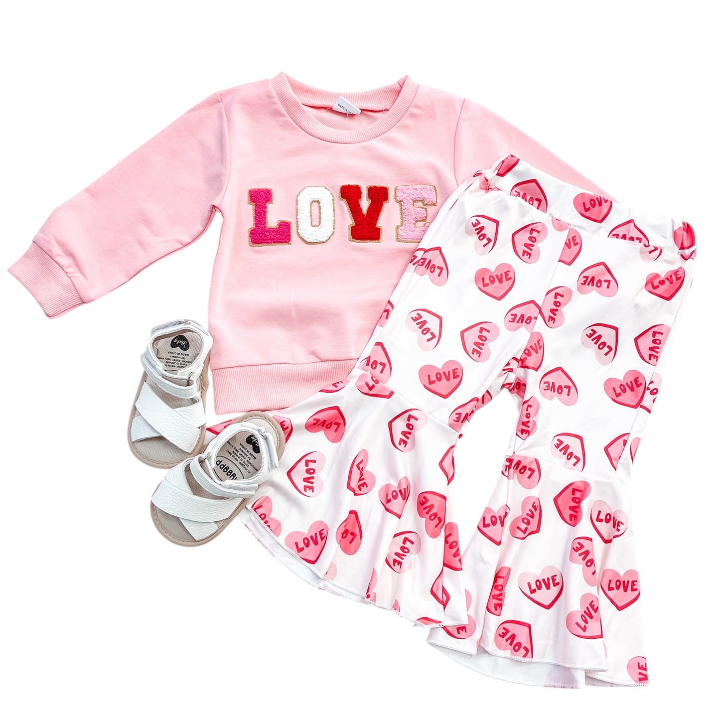 The Tiny Tot Bundles Sweet Love Set, featuring a pink sweatshirt with colorful LOVE letters, white pants with pink heart and LOVE prints, and white sandals for kids, displayed on a white background.