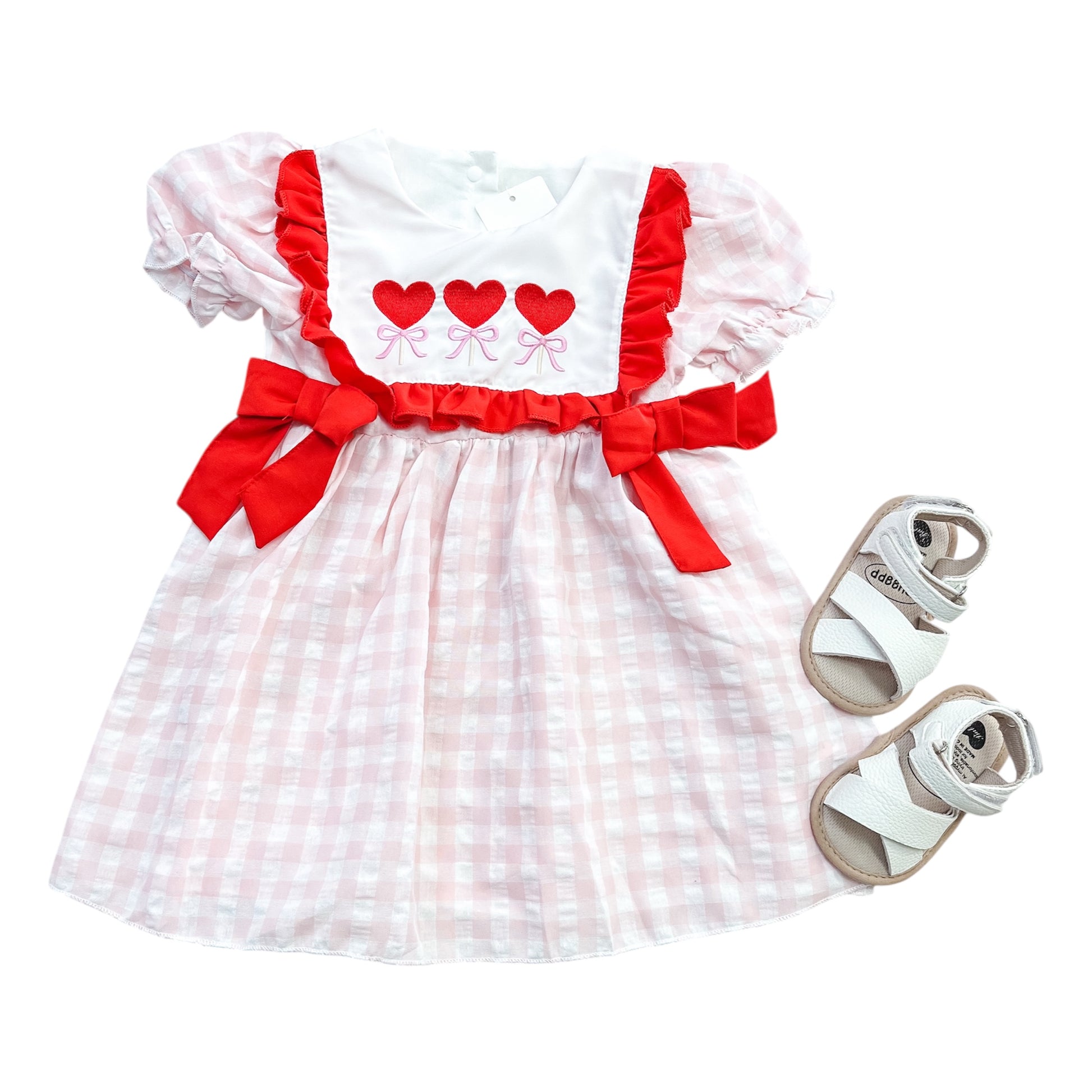 The Sweetheart Trio Dress by Tiny Tot Bundles features pink and white checks, red bows, and three red hearts on the chest, paired perfectly with white toddler sandals.