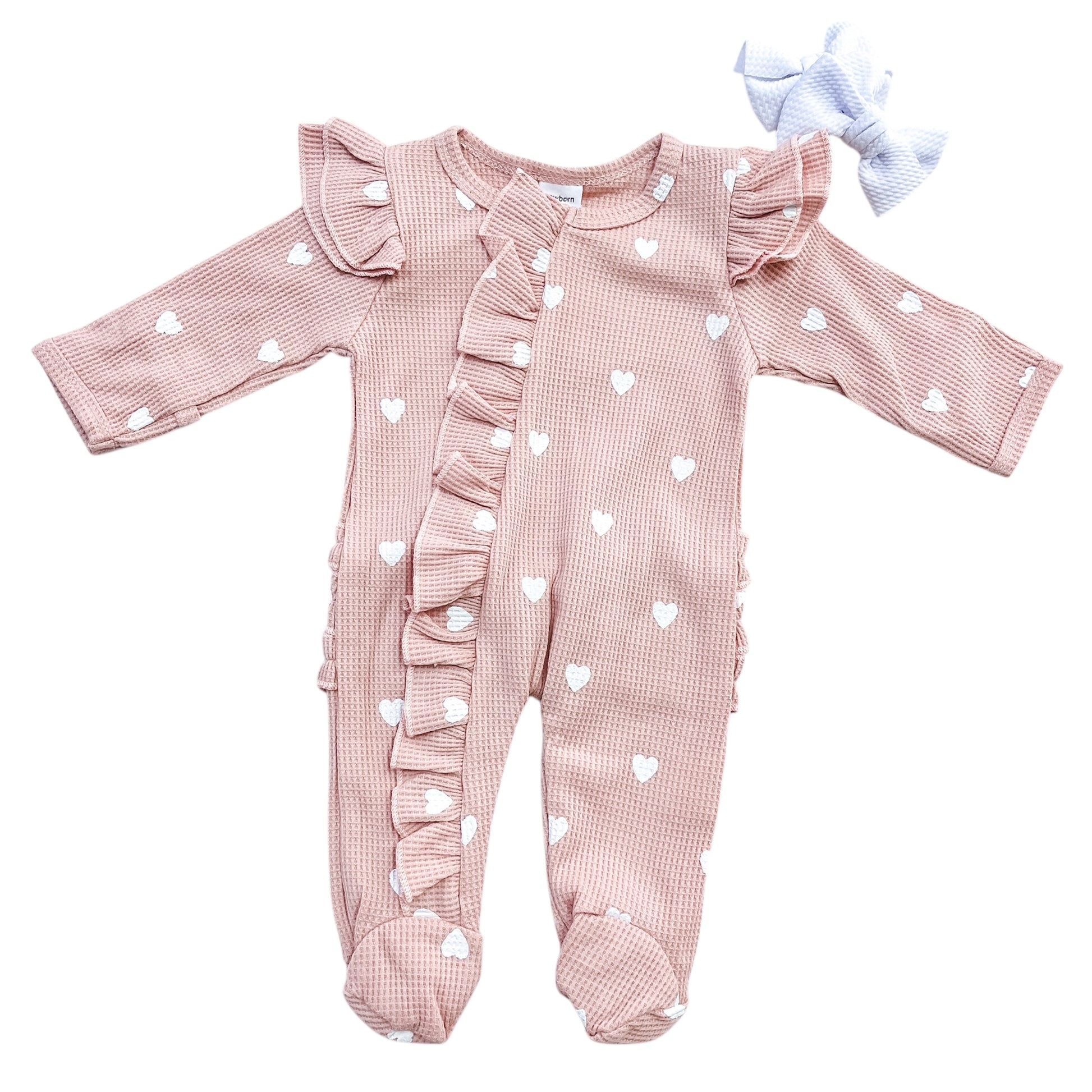 The Tiny Tot Bundles Little Sweetheart Waffle Footie is a pink baby onesie with white hearts, ruffled trim on the front and shoulders, footed legs, and comes with a matching white bow headband.