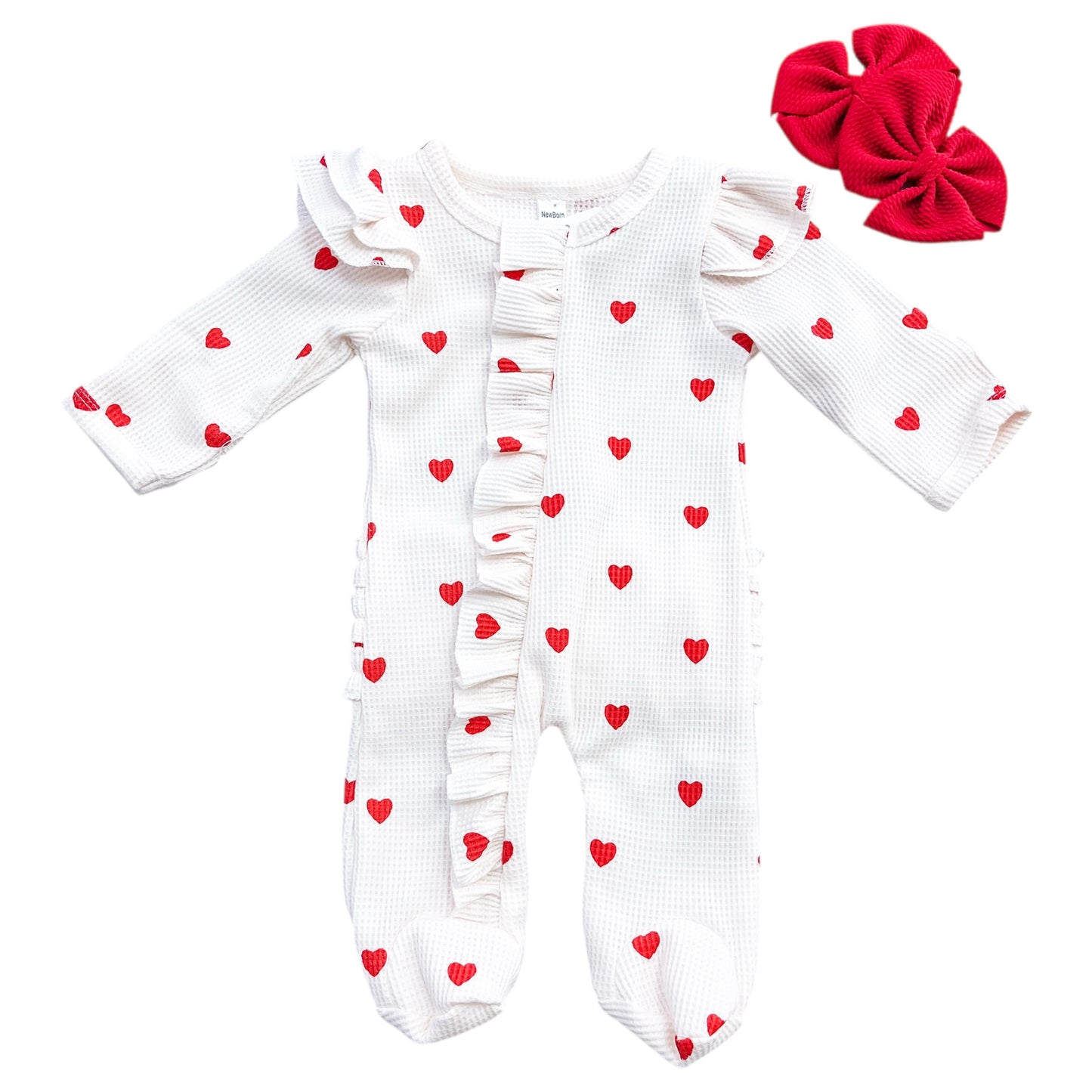 The Little Sweetheart Waffle Footie by Tiny Tot Bundles is a white, long-sleeve baby onesie with red heart prints, ruffled trim on the front and shoulders, plus matching red bow hair accessories shown beside it.