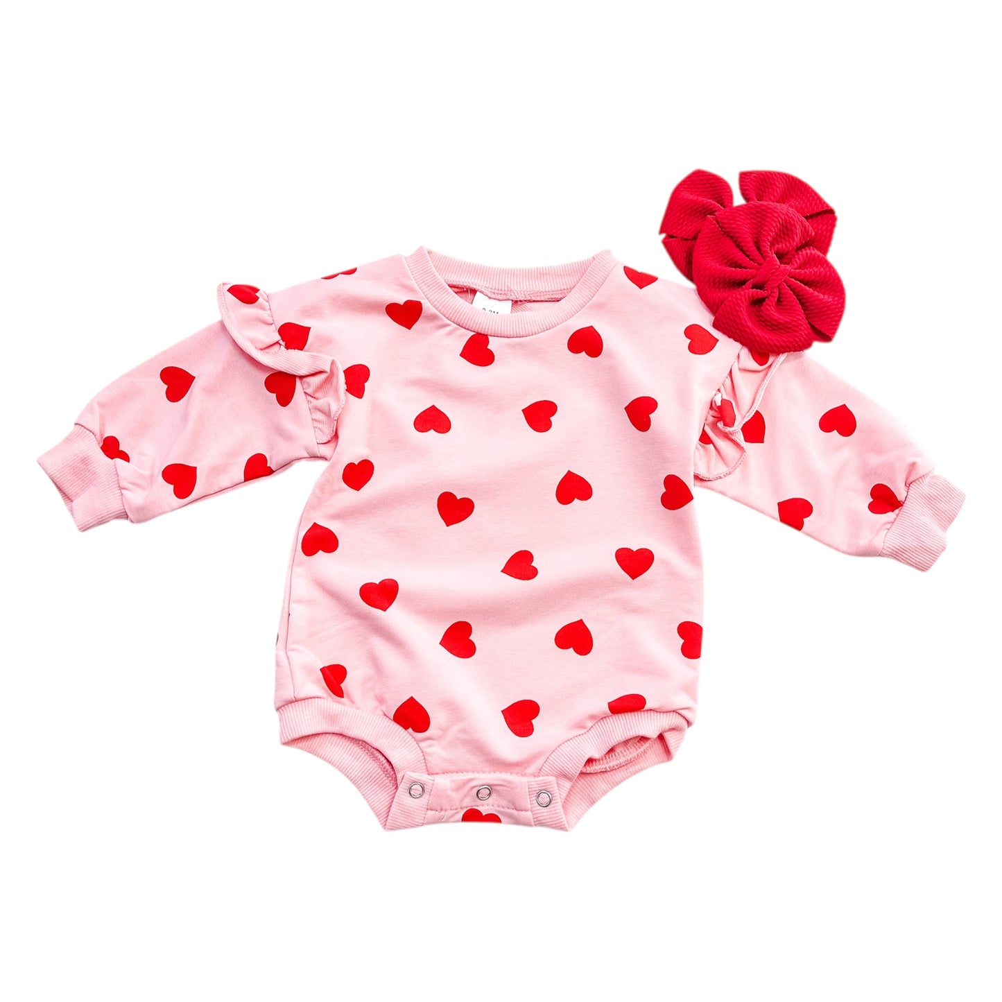 The Tiny Tot Bundles All My Love Bubble is a pink baby romper with long sleeves, red heart prints, ruffled shoulders, and comes with a large red fabric flower accessory.