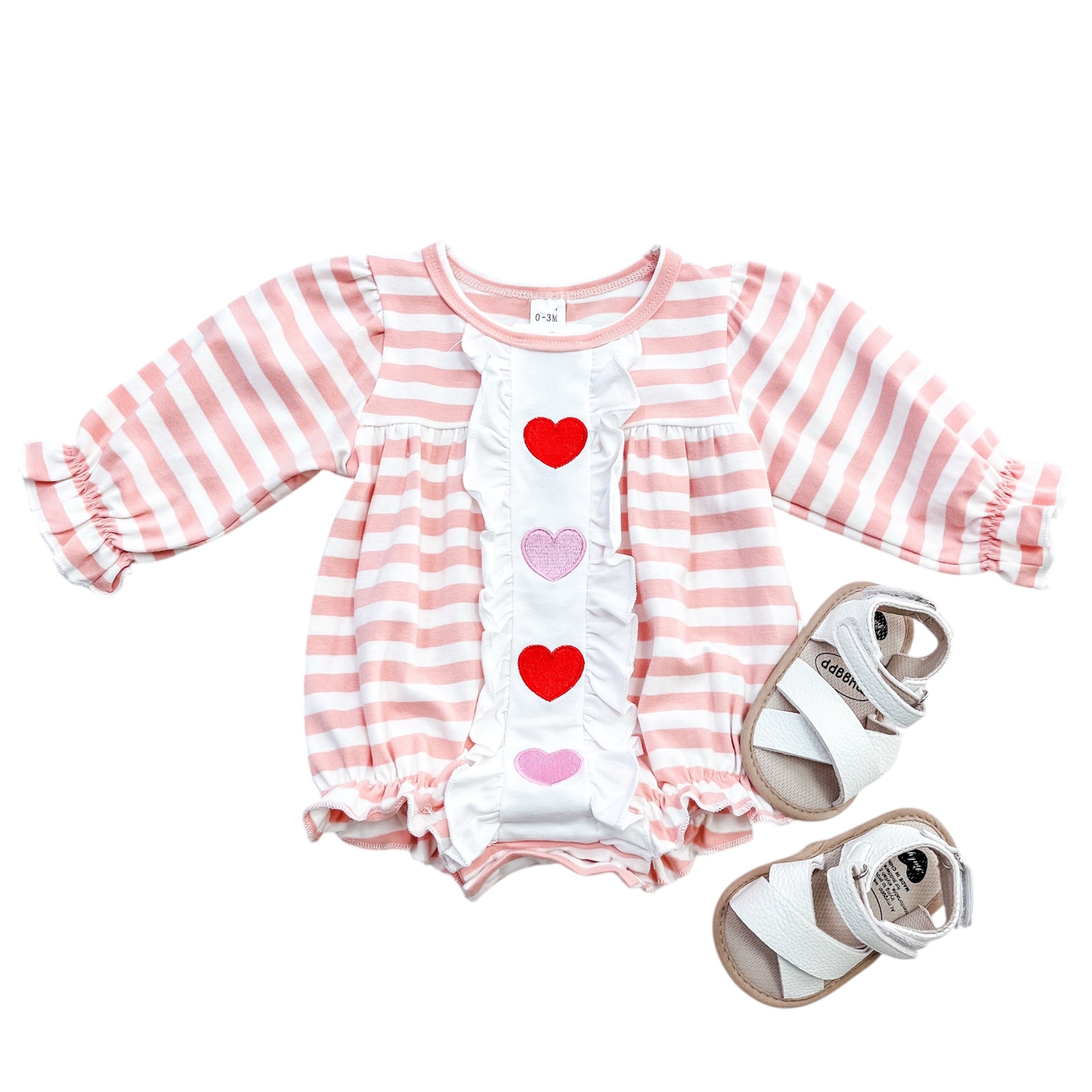 The Rosy Ruffles Hearts Romper by Tiny Tot Bundles features pink and white stripes, long sleeves, ruffled cuffs, and three heart appliqués, shown beside white baby sandals with tan soles and ankle straps.