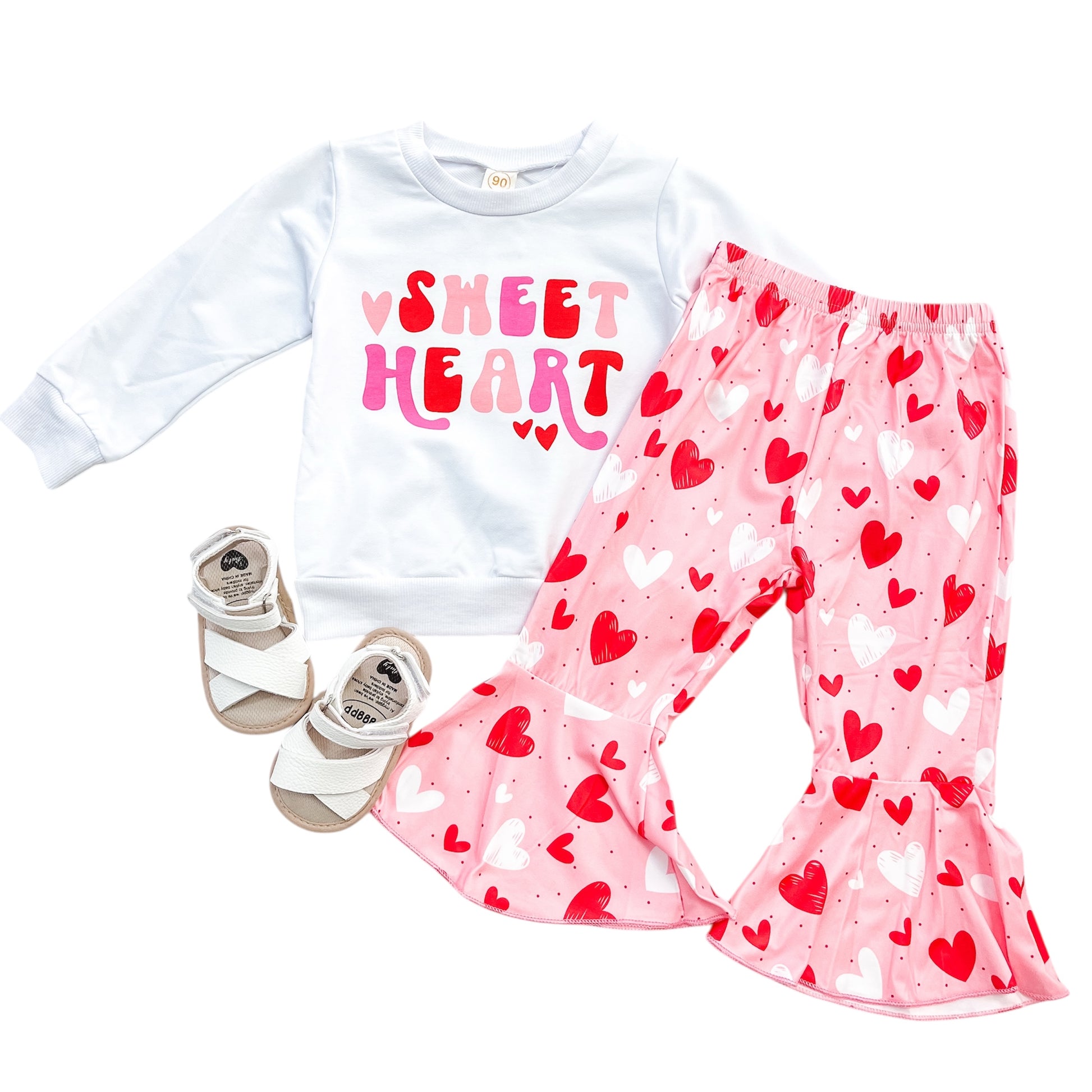 The Sweetheart Vibes Set by Tiny Tot Bundles features a white "Sweet Heart" sweatshirt, pink ruffle pants with red and white hearts, and white sandals—perfect for your child’s adorable style.