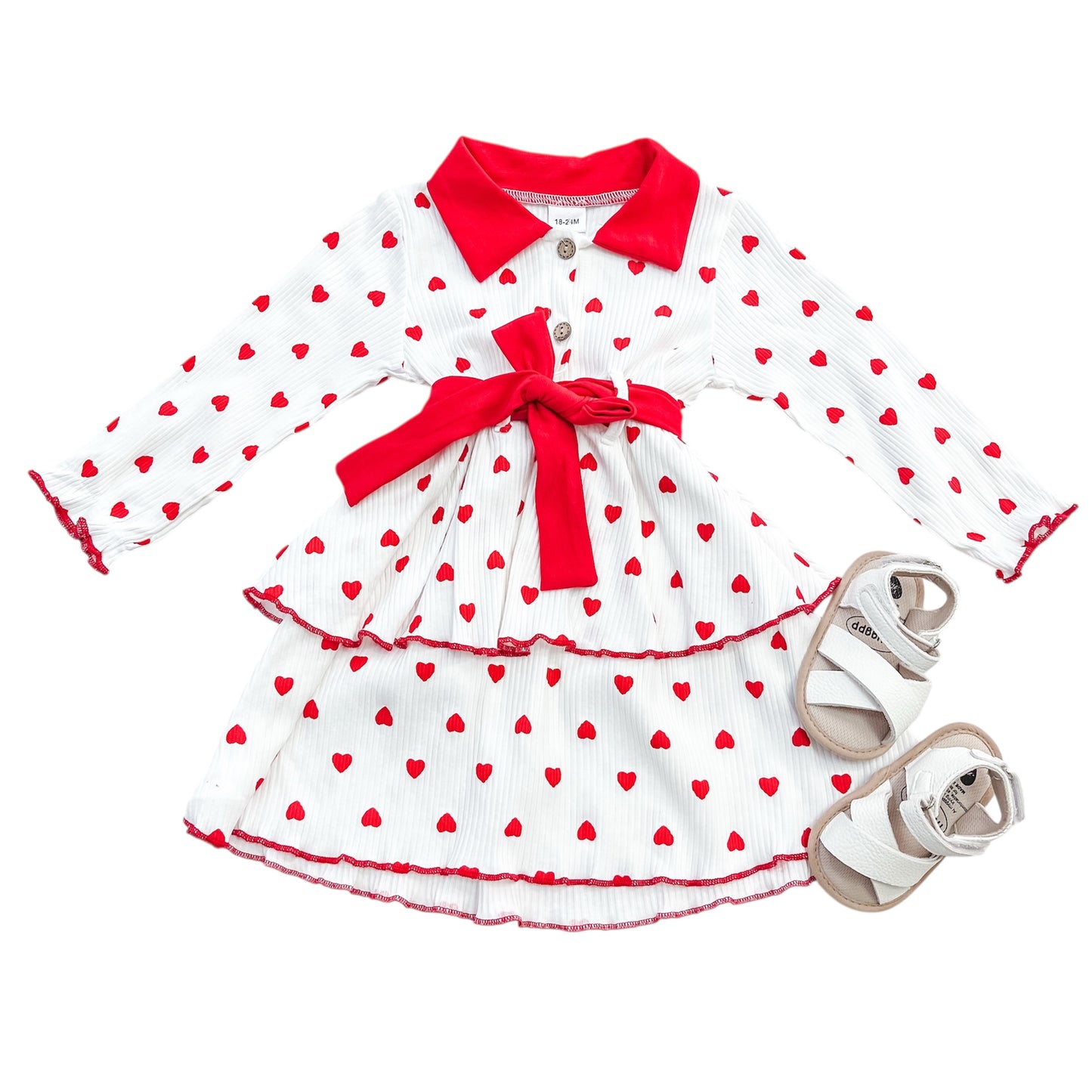 The Scarlet Hearts Tiered Dress by Tiny Tot Bundles features white long sleeves, red heart prints, a red collar, waist bow, and two tiers; pair it with white sandals for a charming look.