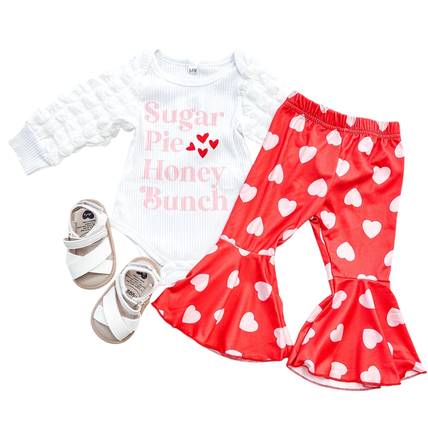 The Sugar Pie Honey Bunch Set by Tiny Tot Bundles includes a white long-sleeve onesie with “Sugar Pie, Honey Bunch” in pink and red, red bell-bottom pants with white hearts, and matching white sandals.