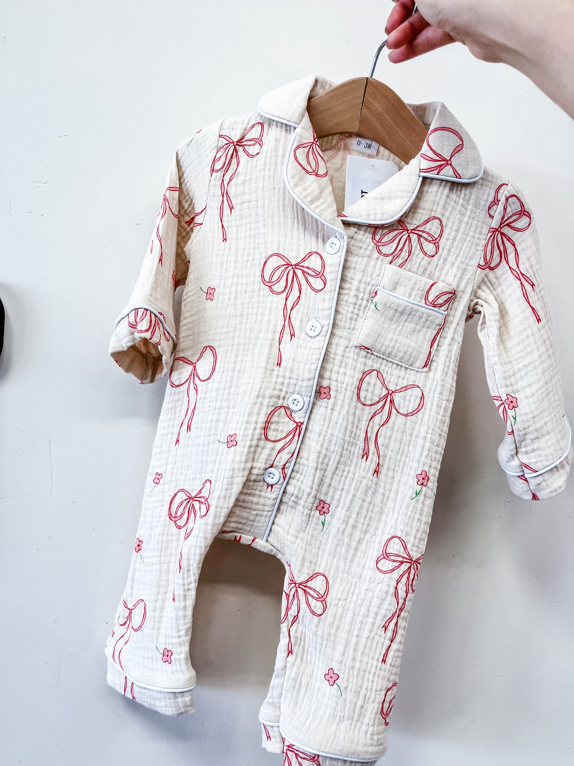 The Tiny Tot Bundles Heirloom Bow Pajama Romper, a cream baby romper with long sleeves, red bow, and pink flower prints, hangs on a wooden hanger held by hand against a plain white background.
