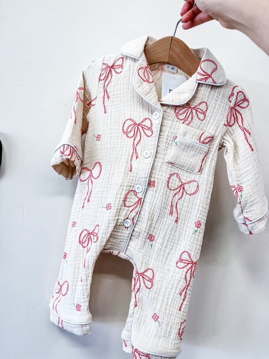 The Tiny Tot Bundles Heirloom Bow Pajama Romper, a cream baby romper with long sleeves, red bow, and pink flower prints, hangs on a wooden hanger held by hand against a plain white background.