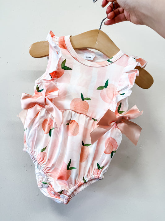 A hand holds the Tiny Tot Bundles Peaches & Cream Bow Bubble, a baby romper with a pink and white checkered pattern, peach prints, ruffled sleeves, and two pink bows at the waist.