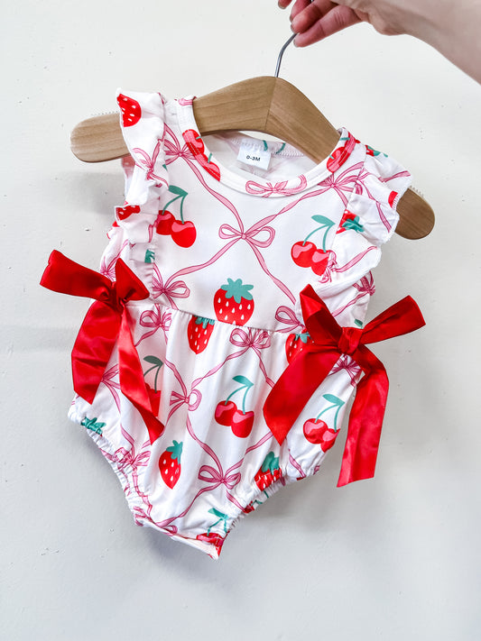 The Berry Sweet Bow Bubble by Tiny Tot Bundles is a white baby romper featuring red strawberry and cherry prints with two large red ribbon bows at the waist, shown on a wooden hanger held by hand.