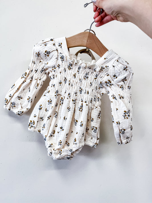 A hand holds a hanger displaying the Tiny Tot Bundles Meadow Whisper Smocked Bubble, a long-sleeved baby romper with small floral print, smocked chest detail, and puffy sleeves against a plain white wall.