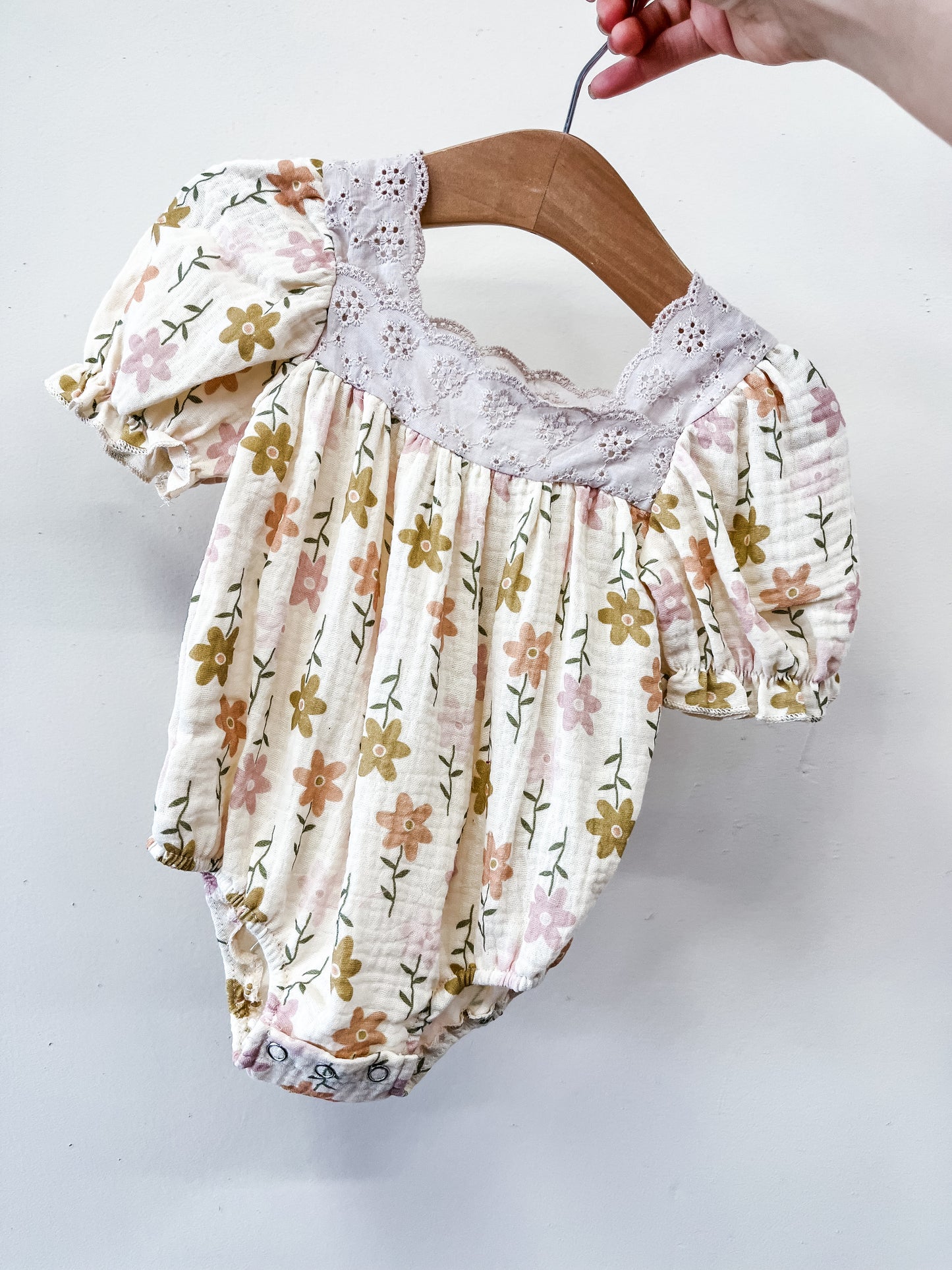 A hand holds Tiny Tot Bundles’ Wildflower Lace Bubble, a baby romper with puff sleeves, yellow and pink floral print, and a white lace collar, displayed on a wooden hanger against a plain white background.
