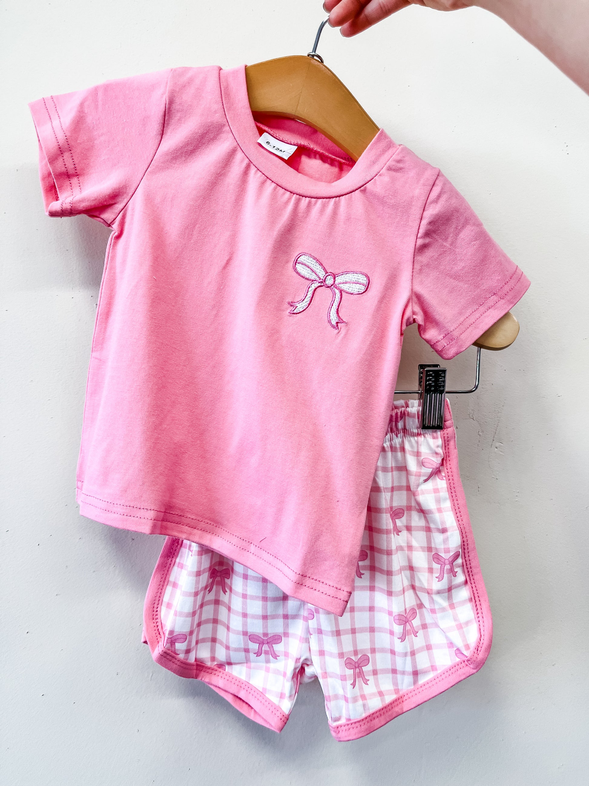 The Pretty Bow Check Set by Tiny Tot Bundles features a pink short-sleeve top with an embroidered bow and matching pink plaid shorts with bow patterns, displayed on a hanger against a white background.