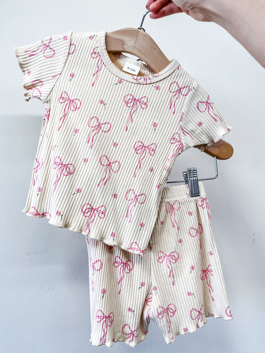 A hand holds the Tiny Tot Bundles Heirloom Bow Lounge Set—a matching toddler outfit on a hanger with a cream background, pink bows, tiny flowers, and ribbed texture. The set includes a short-sleeve shirt and shorts.