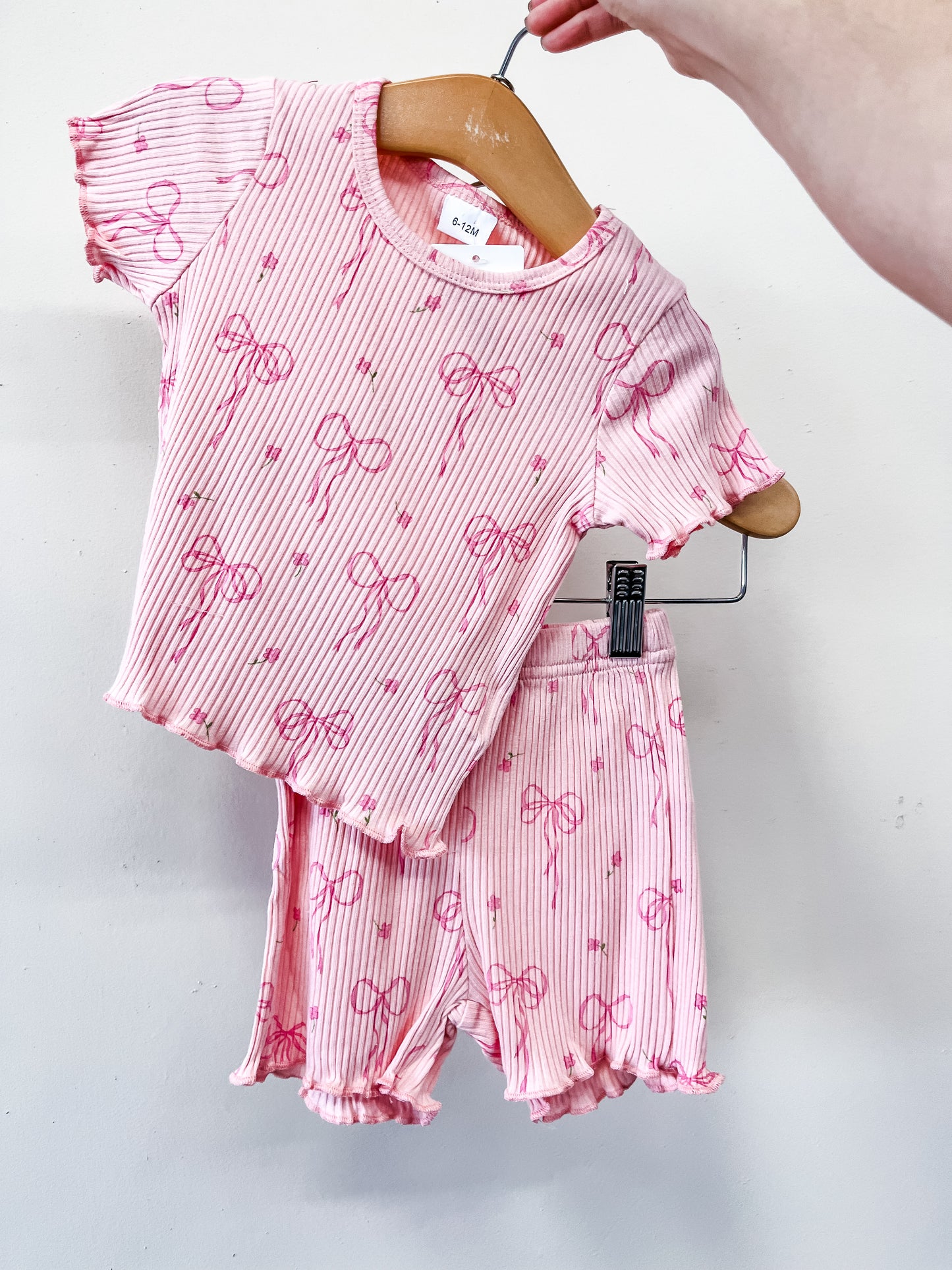 A hand holds a hanger displaying the Blushing Bows Lounge Set by Tiny Tot Bundles, a matching pink toddler outfit with short sleeves and shorts, featuring a charming pattern of small bows and stripes.