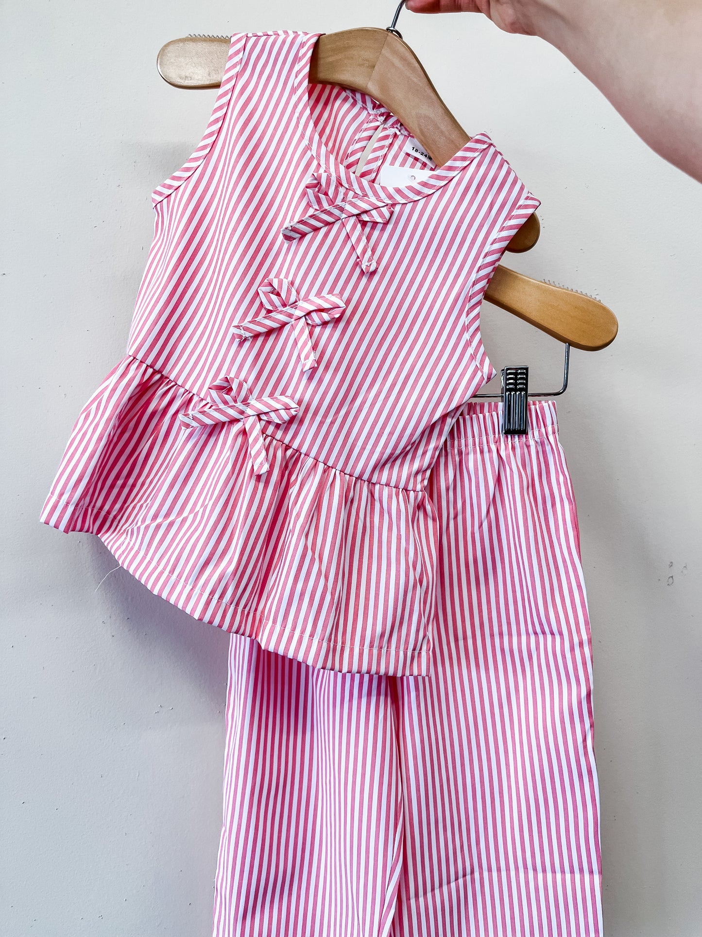 The Sweet Stripe Bow Set by Tiny Tot Bundles features a pink-and-white striped sleeveless top with ruffle accents and matching pants, displayed on wooden hangers against a simple light background.