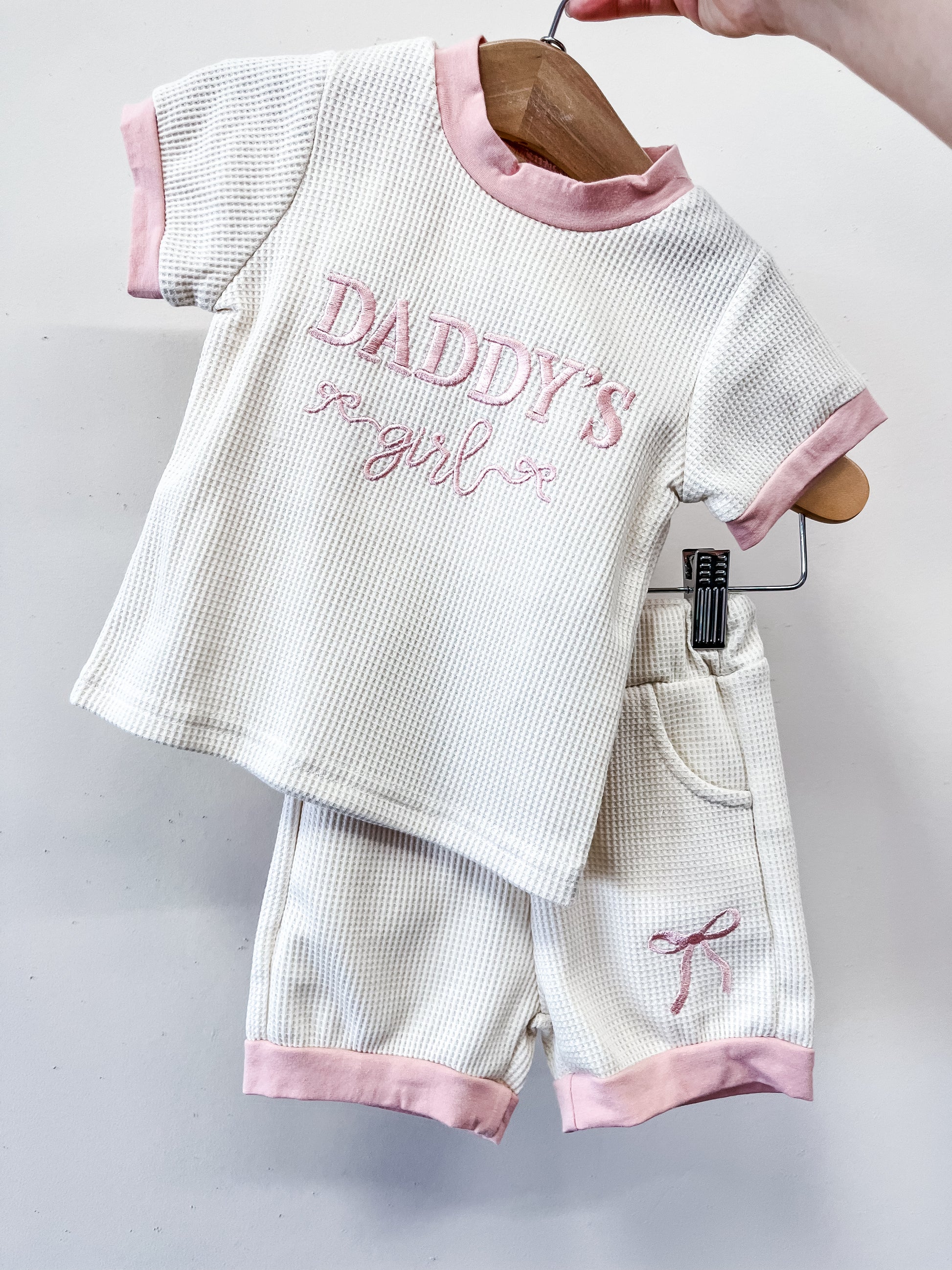 The Daddy's Girl Waffle Lounge Set by Tiny Tot Bundles is a white toddler outfit with pink trim, including a short-sleeve top that says “Daddy’s girl” in pink script and matching shorts with a pink bow, shown on a wooden hanger.