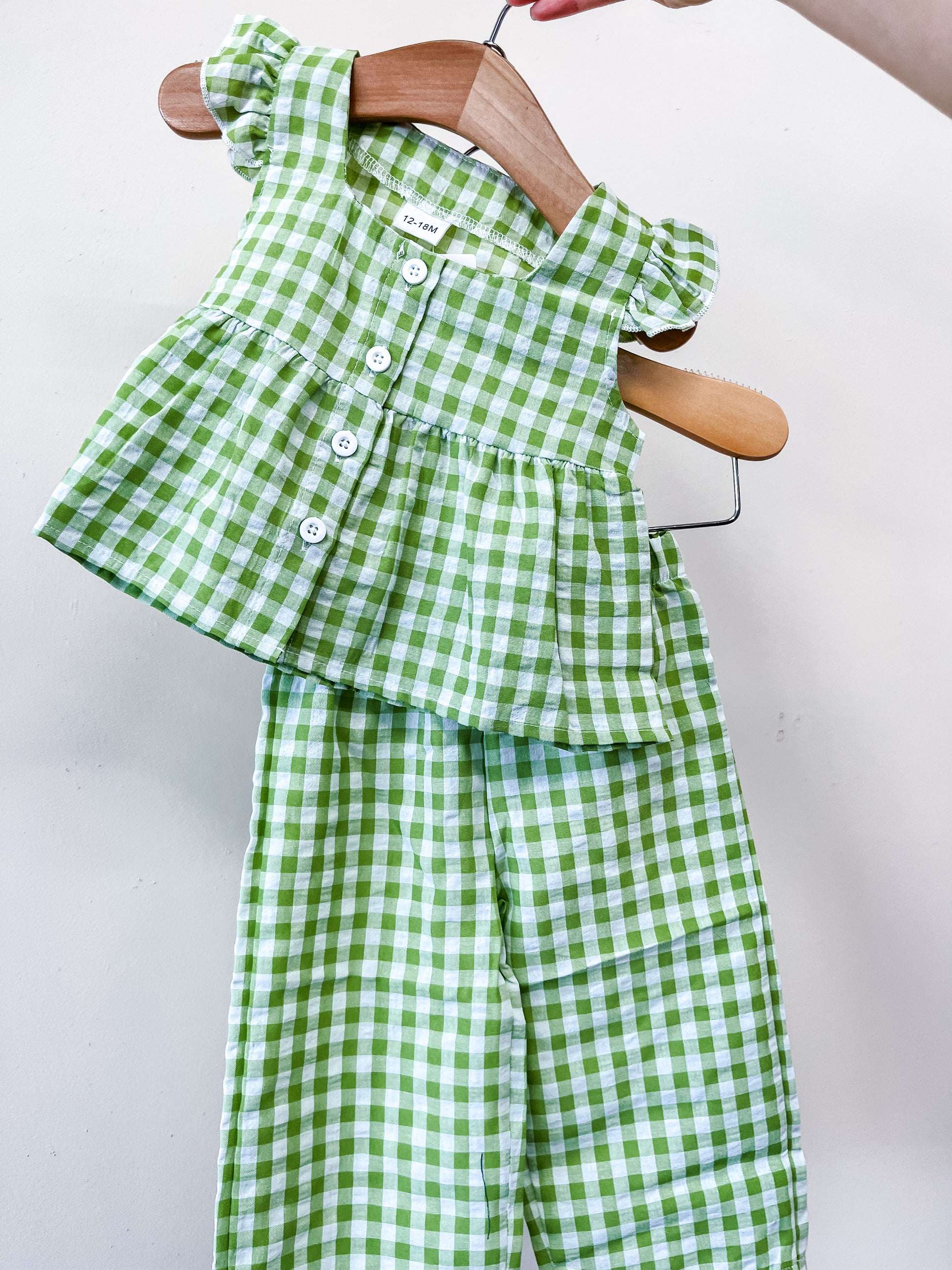 The Sweet Pea Gingham Set by Tiny Tot Bundles features a green and white checkered sleeveless top with ruffled shoulders and white buttons, paired with matching pants, shown on a wooden hanger against a plain background.