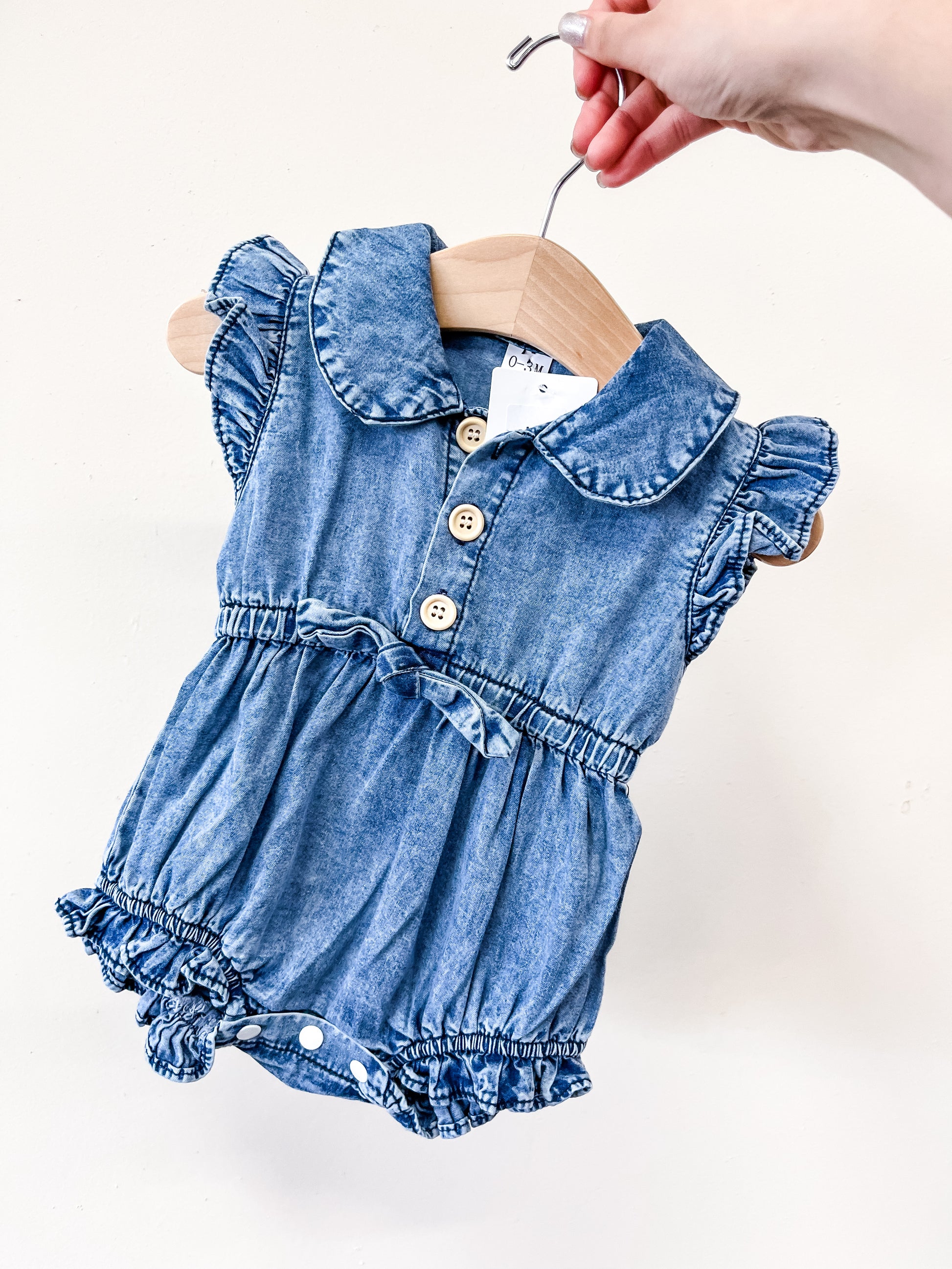 A hand holds a hanger with the Tiny Tot Bundles Denim Ruffle Romper—featuring blue denim, ruffled sleeves, a Peter Pan collar, wooden buttons, waist bow, and elasticized leg openings.