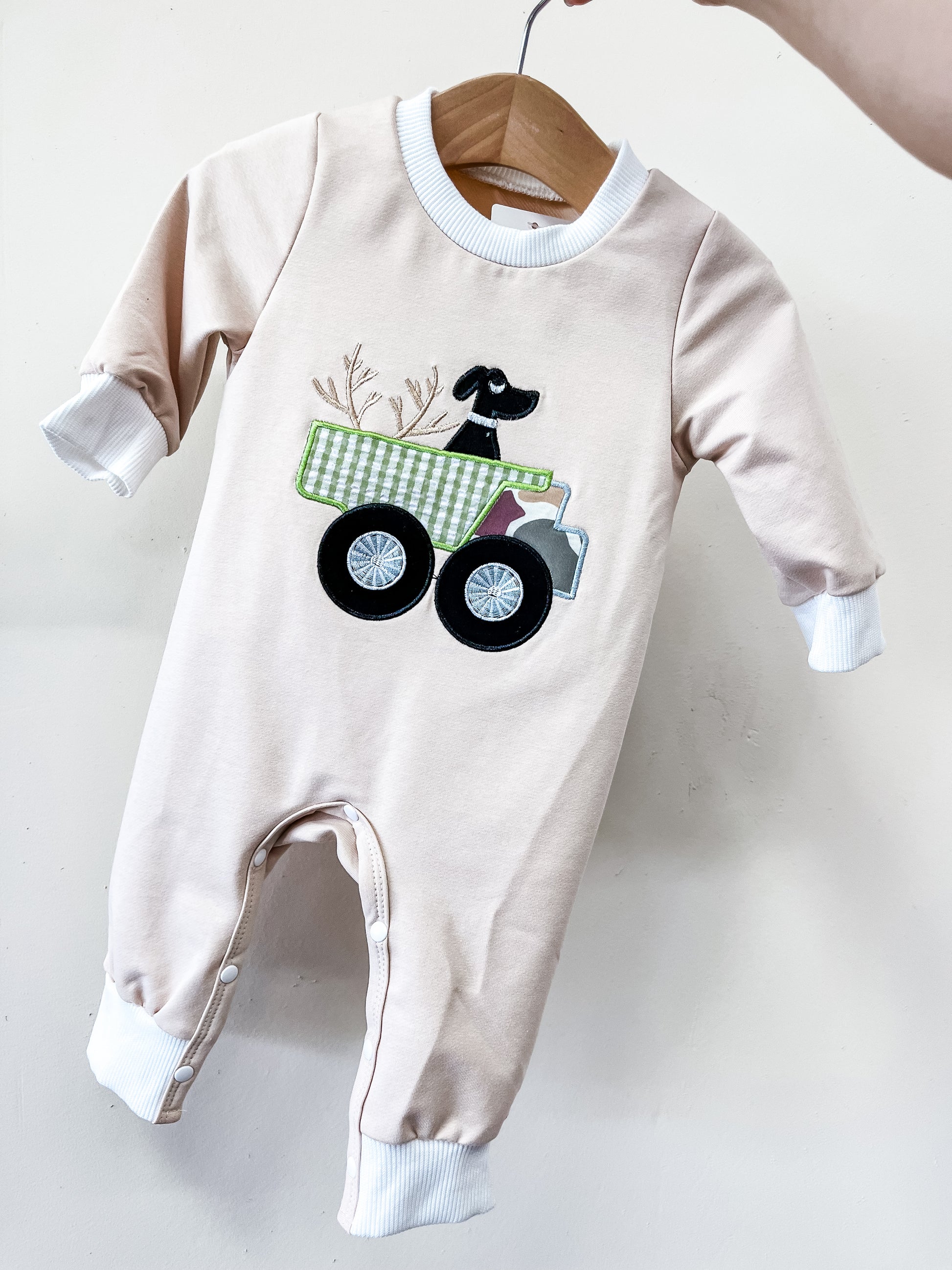 The Little Retriever Wagon Footie by Tiny Tot Bundles is a light pink baby romper with white cuffs, featuring a black dog in a green checkered truck applique and branches in back, displayed on a wooden hanger.