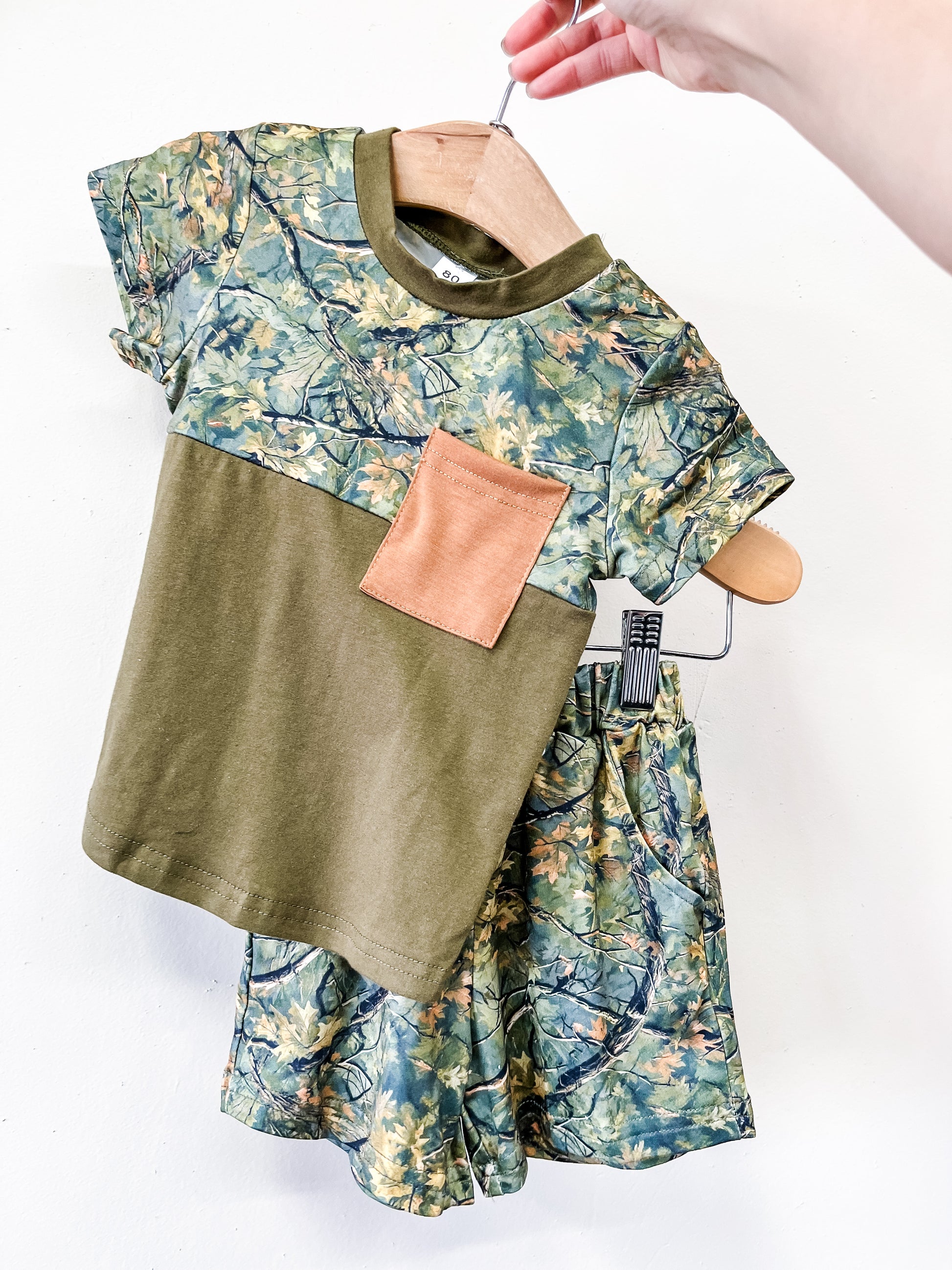 A hand holds the Little Woods Short Set by Tiny Tot Bundles: a child's green camo shirt with a brown chest pocket and matching leafy camouflage shorts, shown on a hanger against a plain white background.