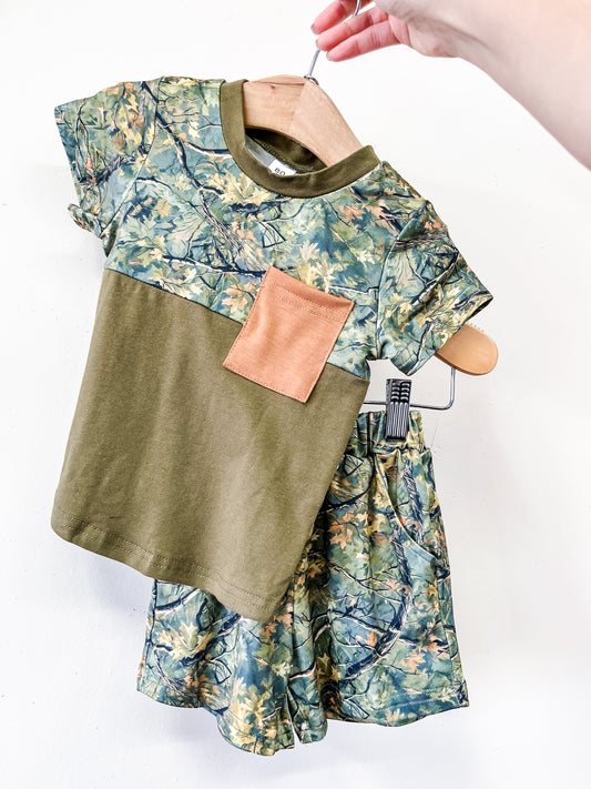 A hand holds the Little Woods Short Set by Tiny Tot Bundles: a child's green camo shirt with a brown chest pocket and matching leafy camouflage shorts, shown on a hanger against a plain white background.