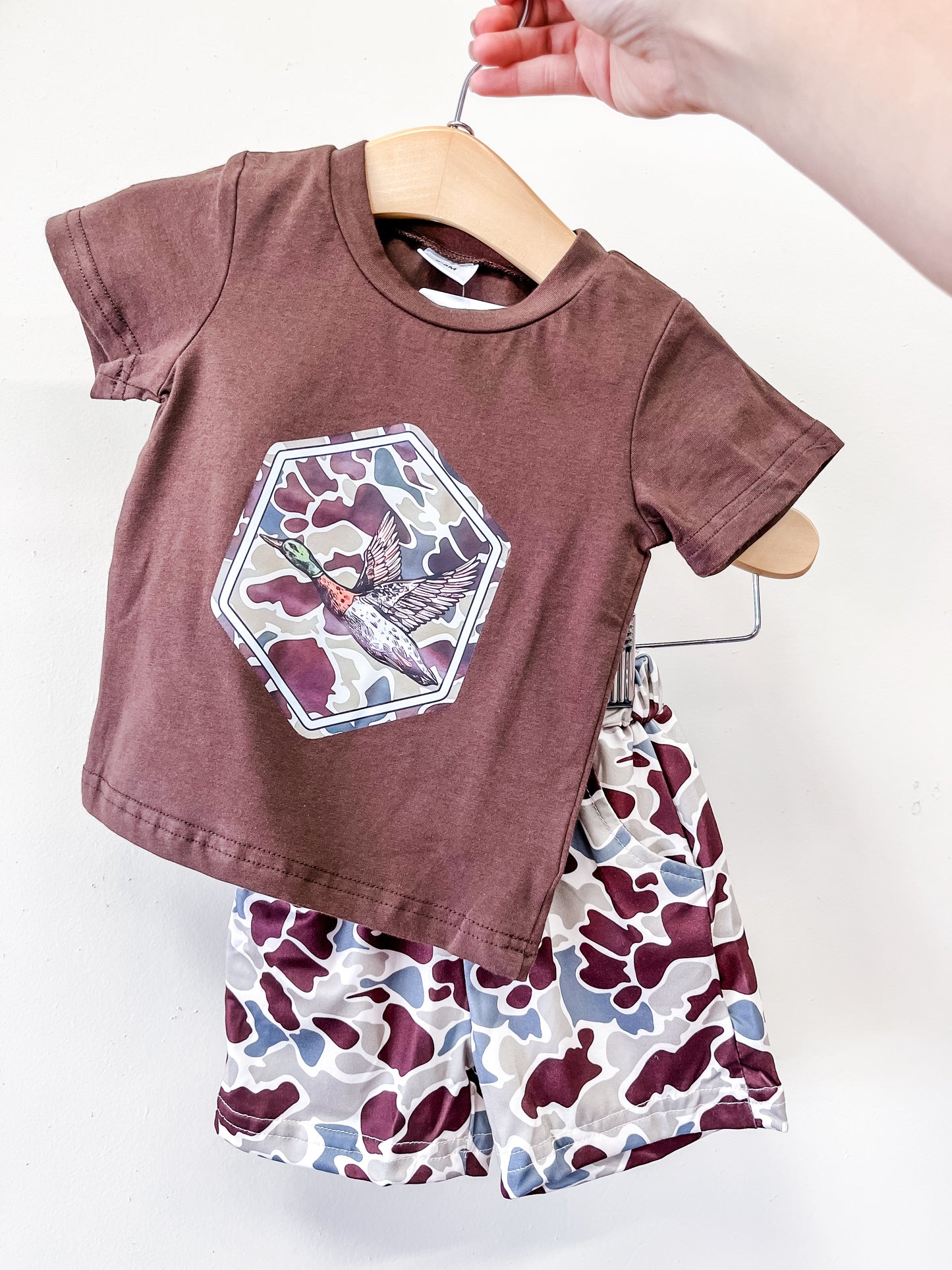 A hand holds a hanger displaying the Tiny Tot Bundles Duck Hunter Short Set: a brown short-sleeve t-shirt with bird and geometric designs, paired with brown, white, and gray camo-patterned shorts for children.