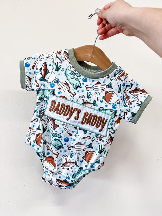 A hand holds the Tiny Tot Bundles Daddy's Buddy Fishing Bubble, a baby onesie with fishing-themed designs, "Daddy's Buddy" on the front pocket, and green trim on the sleeves and neck.