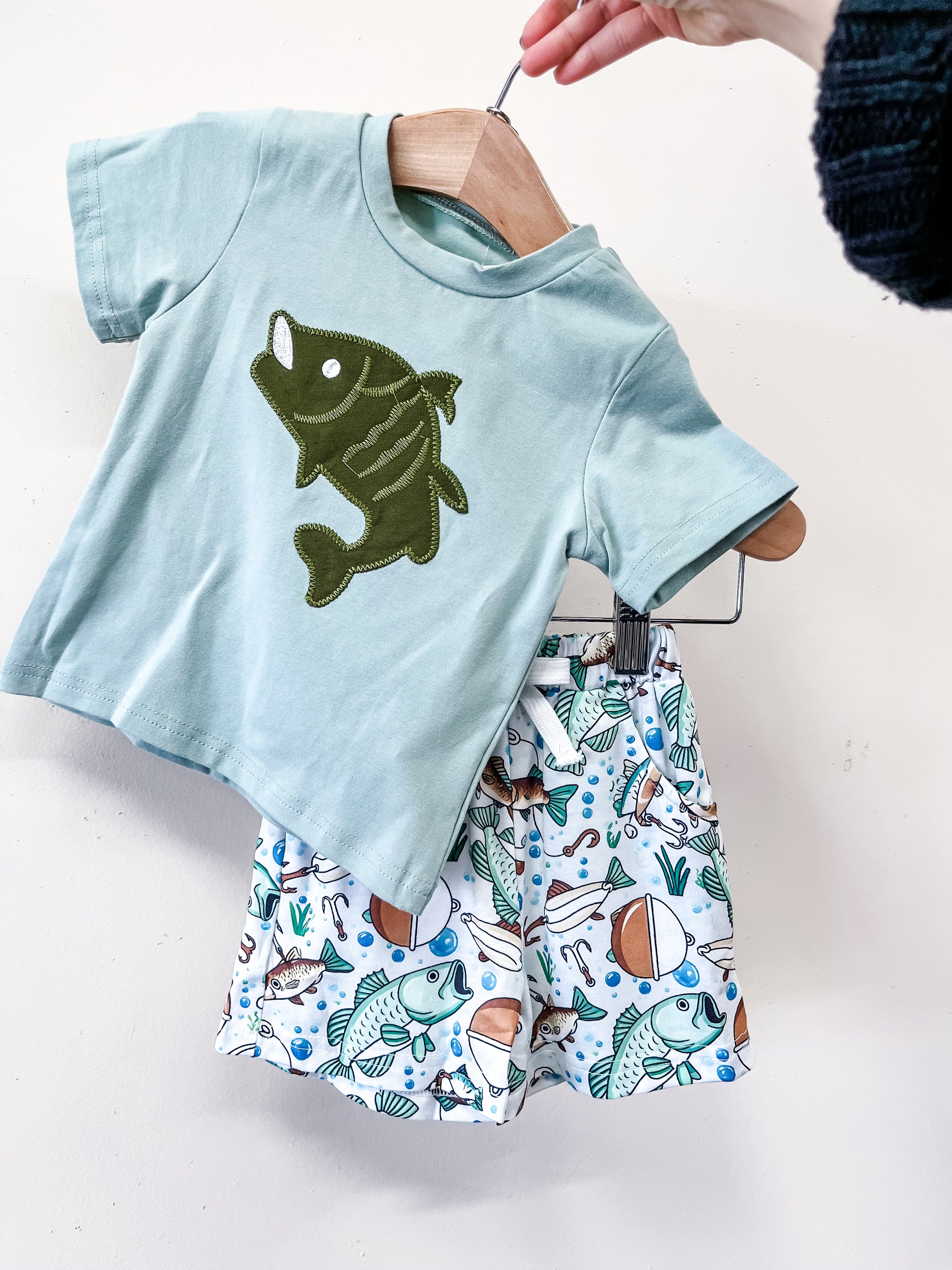A hand displays the Little Catch Short Set by Tiny Tot Bundles, featuring a light blue tee with a large green fish and coordinating shorts with colorful fish and fishing-themed patterns.