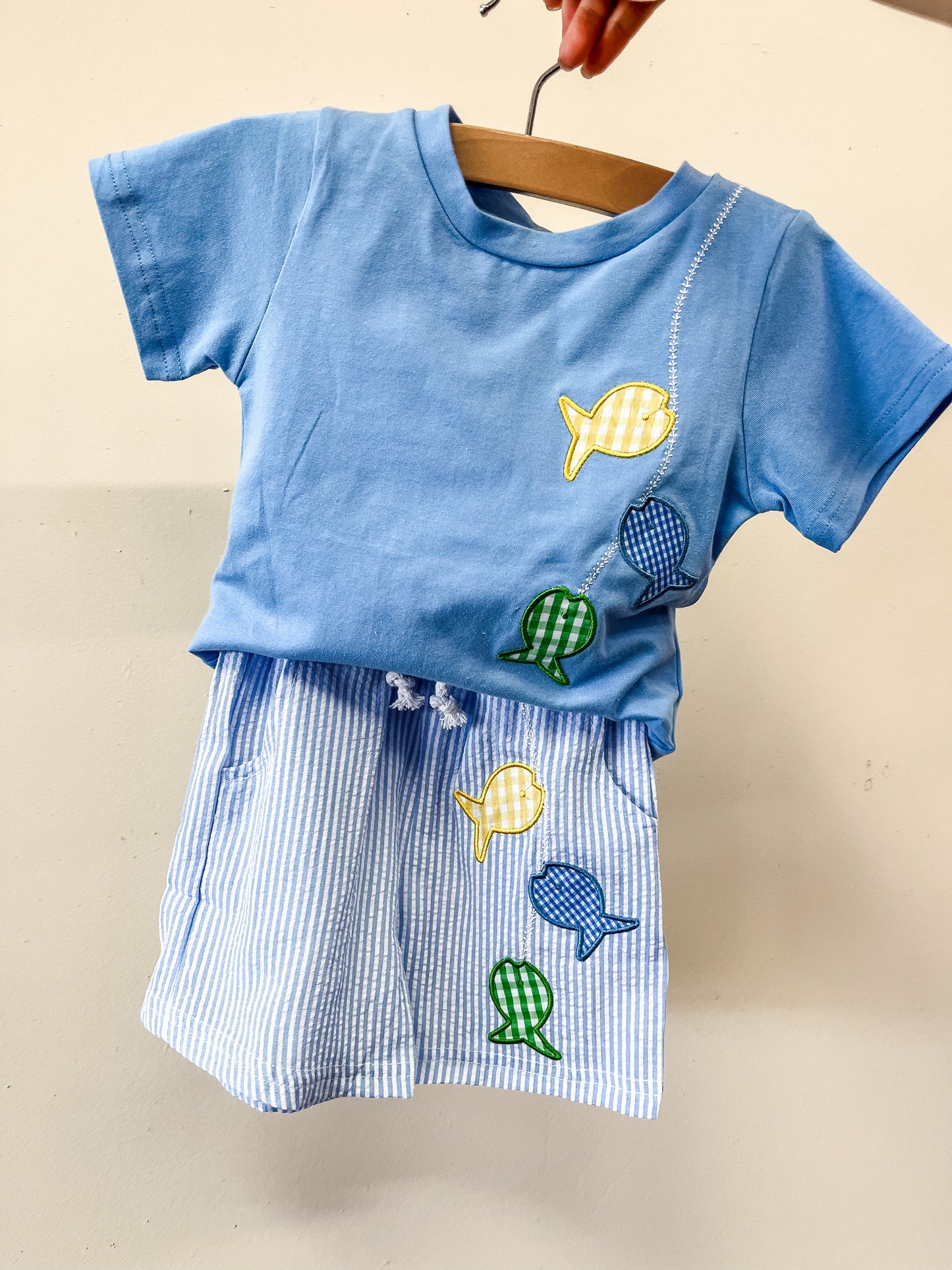 The Gone Fishing Short Set by Tiny Tot Bundles features a blue toddler t-shirt and striped shorts on a hanger, both adorned with embroidered fish in yellow, green, and blue.