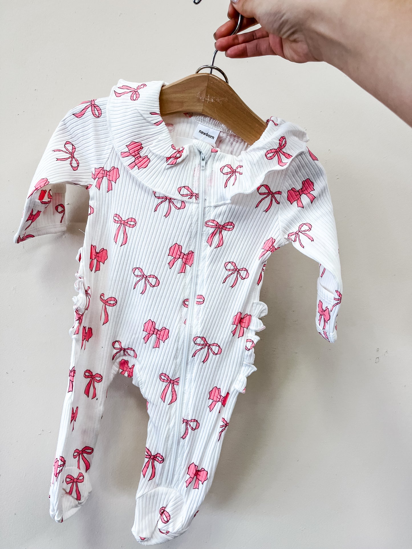 A person holds up the Tiny Tot Bundles Bow Darling Ruffle Footie, a white baby onesie with pink bows, ruffled collar, and ruffle details along the zipper and sides, displayed on a wooden hanger against a plain background.