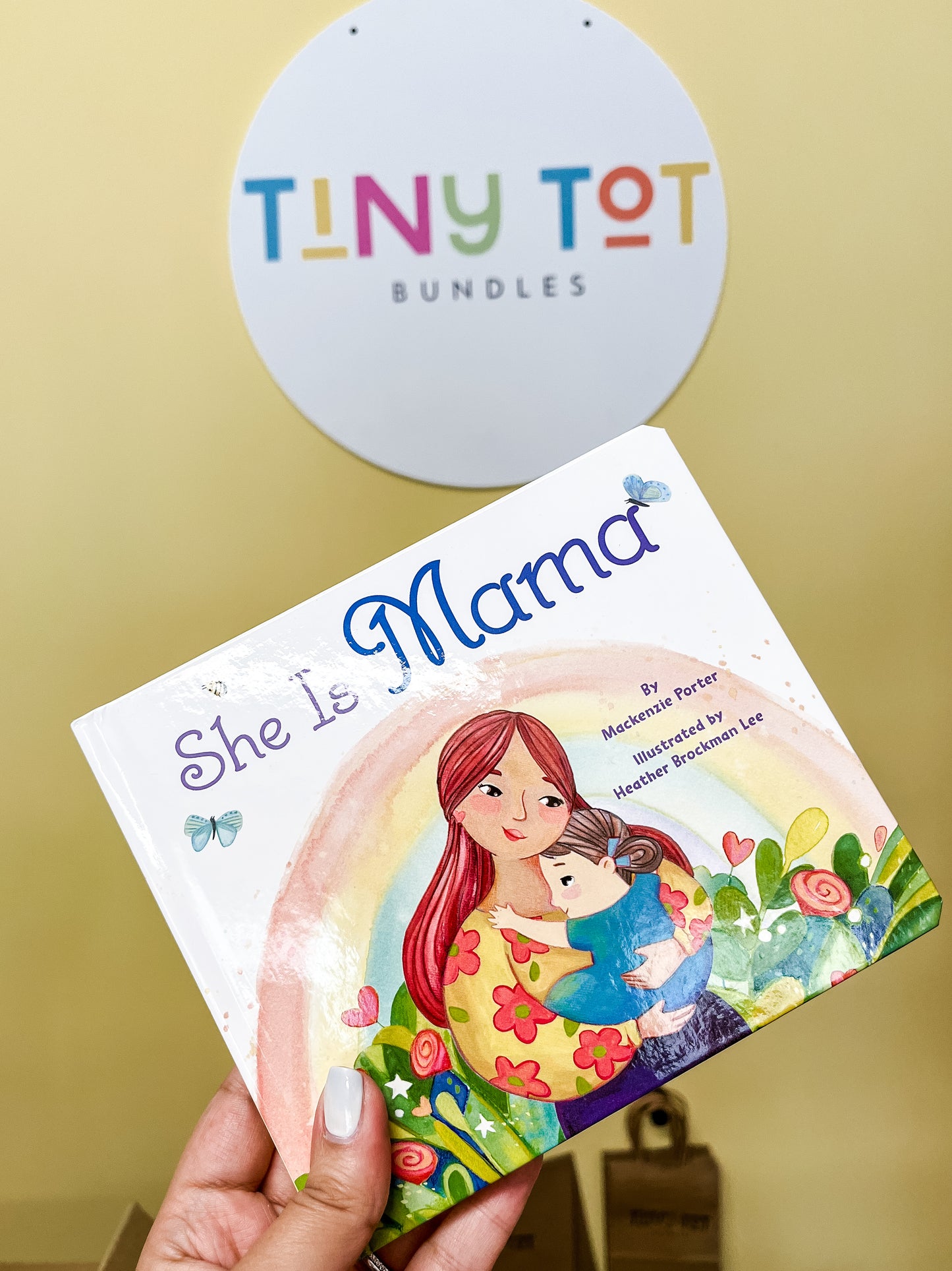 A hand holds the She Is Mama Book by Tiny Tot Bundles in front of a wall sign reading Tiny Tot Bundles. The cover shows an illustration of a woman hugging a child, surrounded by flowers and colorful patterns.