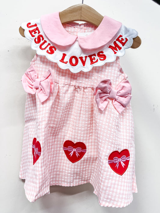 The Jesus Loves Me Sweetheart Dress by Tiny Tot Bundles is a pink and white checkered baby dress with large side bows, red heart patches, and a white collar featuring “JESUS LOVES ME” in bold red letters. Displayed on a wooden hanger against a white background.