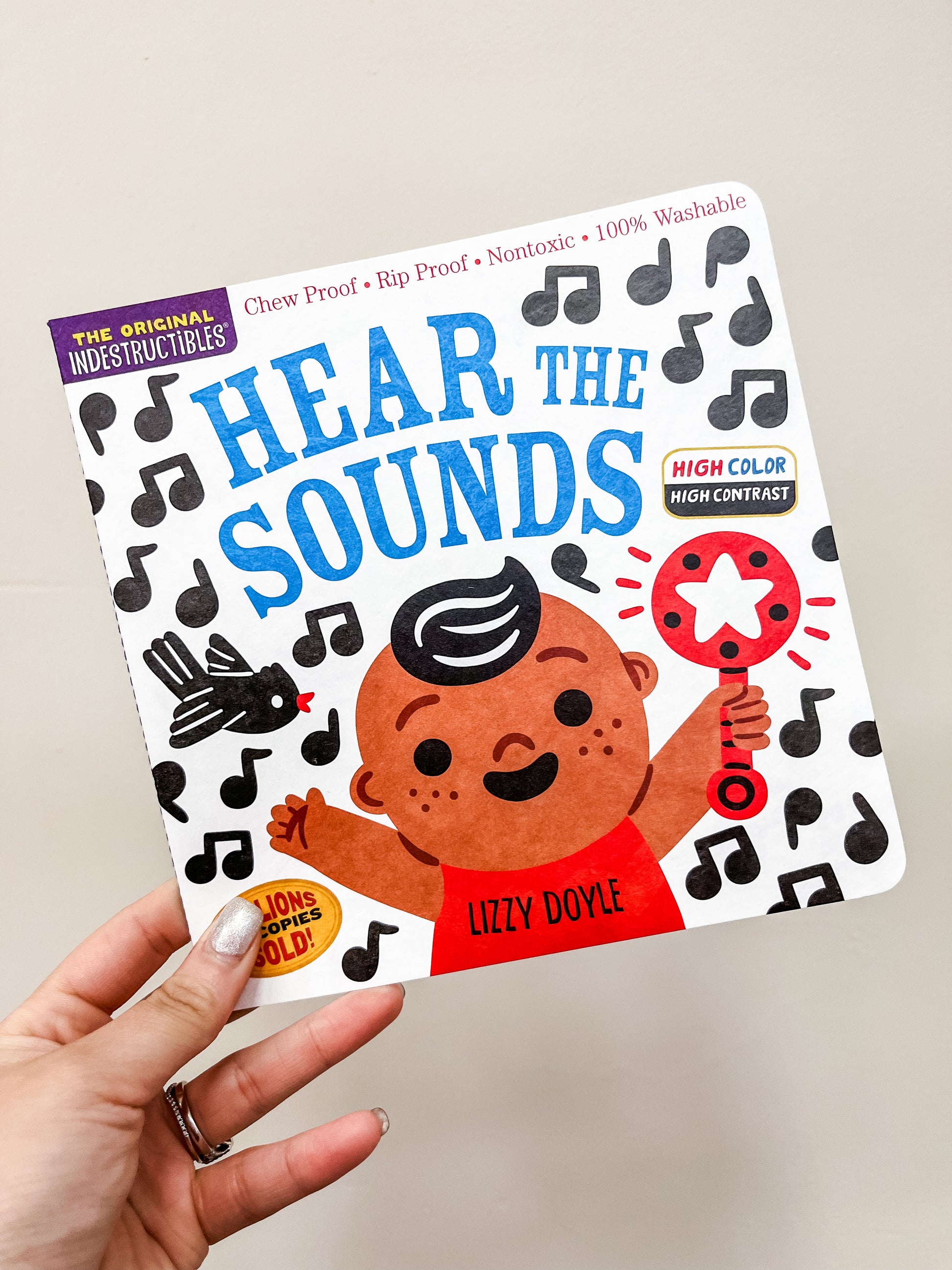 A hand holds Hear The Sounds Book by Hachette Book Group, featuring a smiling child with a rattle, musical notes, and a black bird on the cover. The book is chew proof and nontoxic.