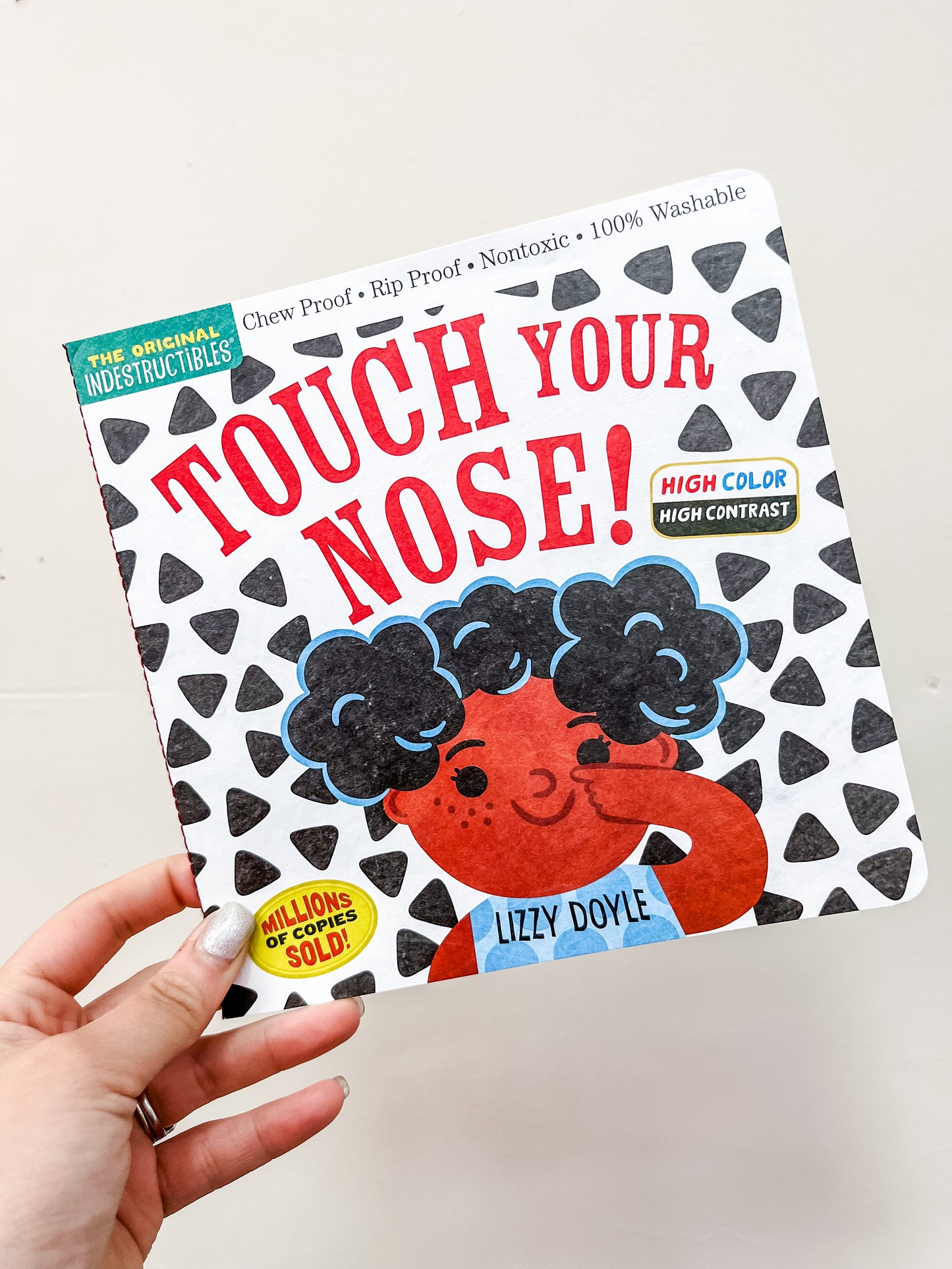 A hand holds the Touch Your Nose Book by Hachette Book Group, showing an illustrated child with curly hair touching their nose on a white cover decorated with black triangle patterns.