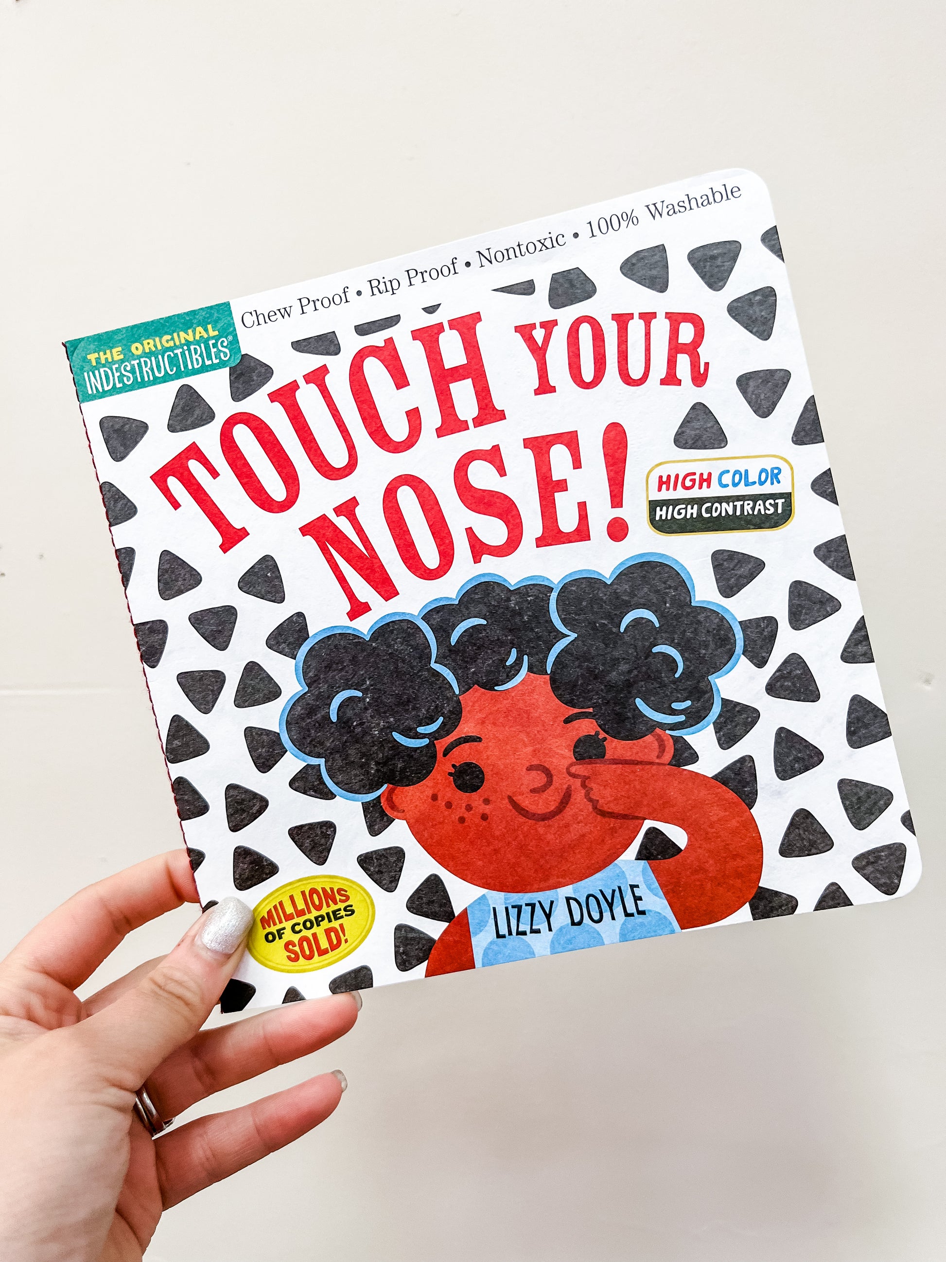 A hand holds the Touch Your Nose Book by Hachette Book Group, showing an illustrated child with curly hair touching their nose on a white cover decorated with black triangle patterns.