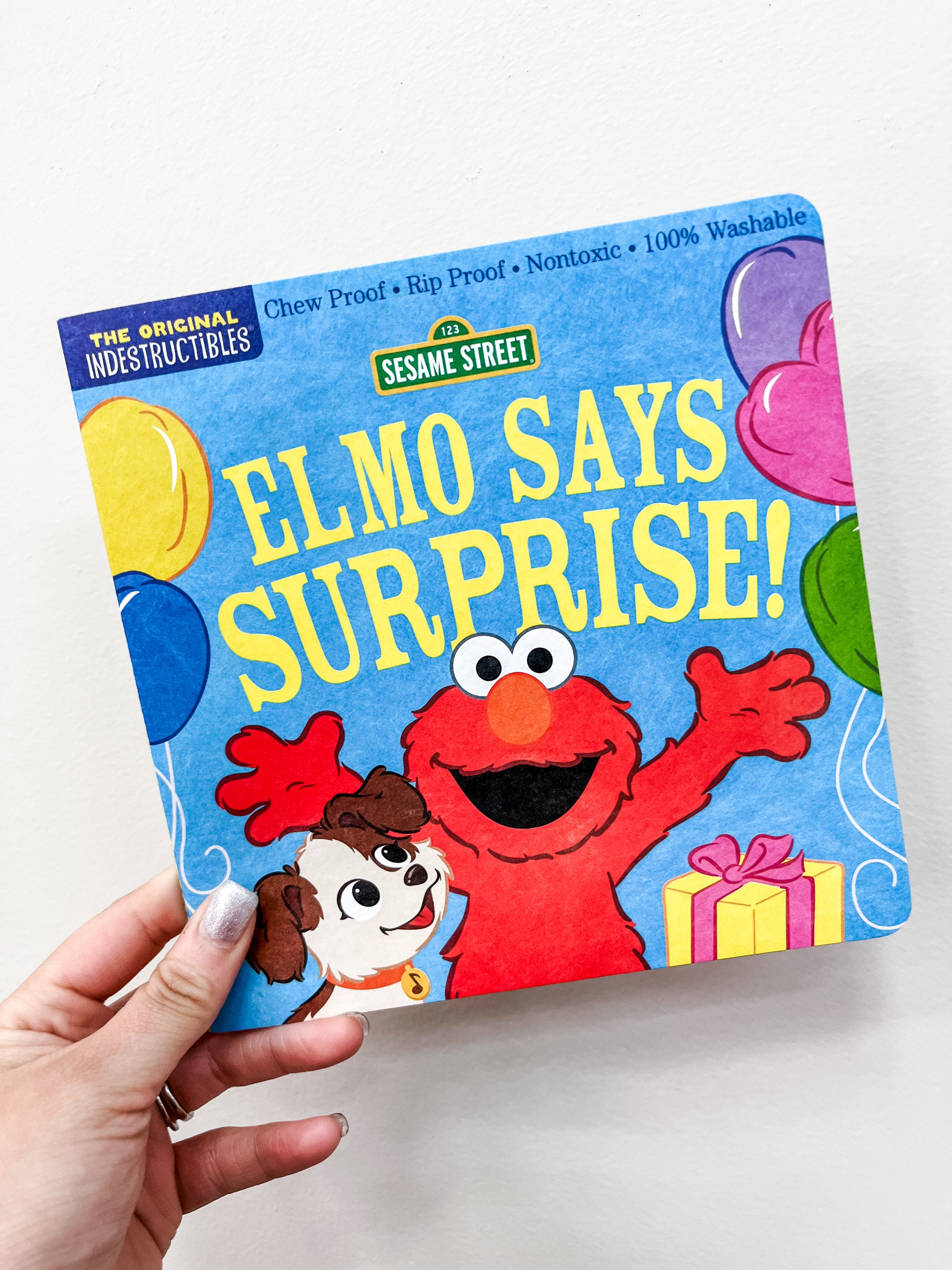 A hand holds the Elmo Says Surprise Book by Hachette Book Group, featuring Elmo with raised arms, a puppy, balloons, and a gift on a playful blue cover illustrated for children.