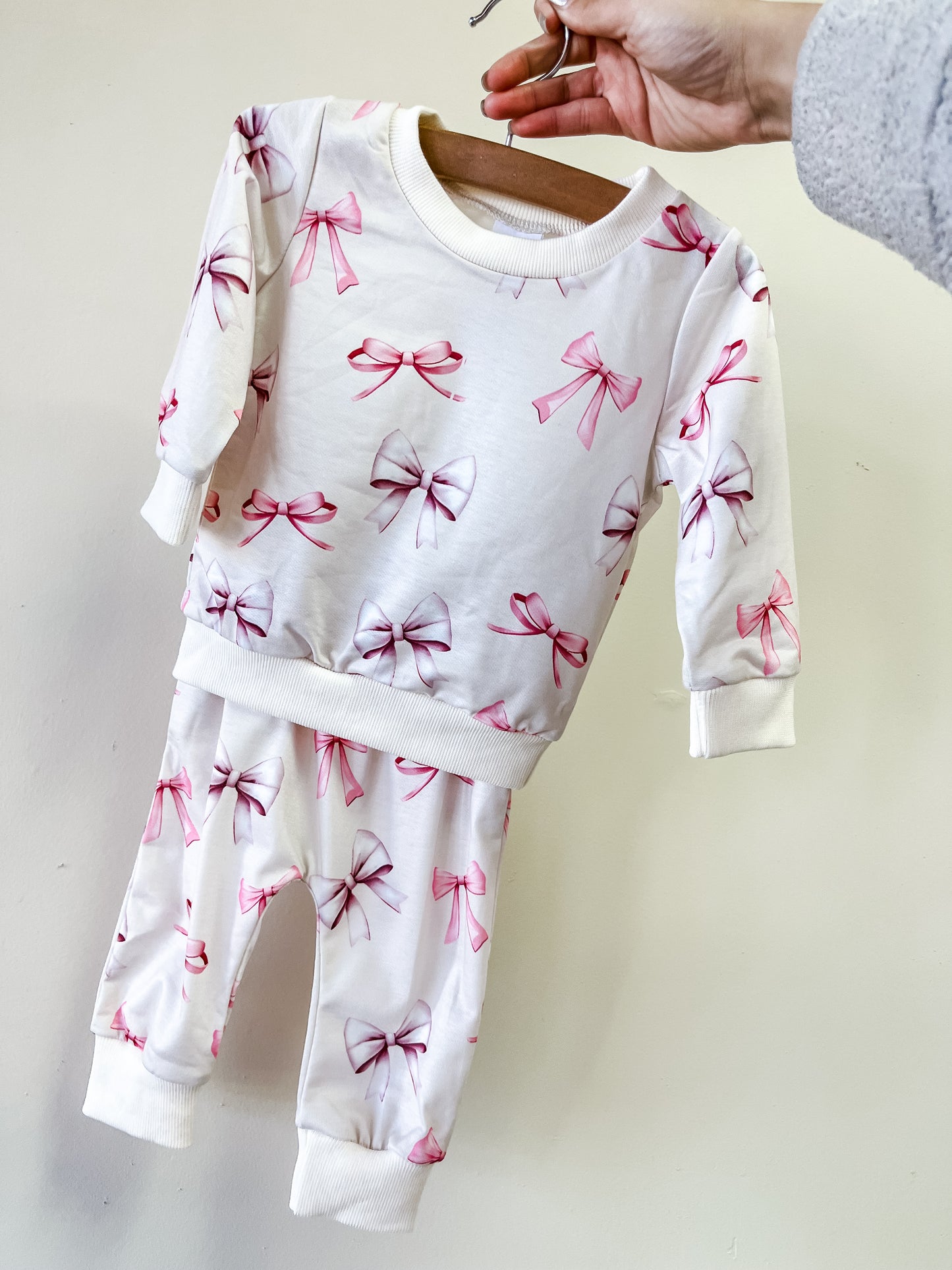 A person holds up the Tiny Tot Bundles Vintage Bow lounge Set, a white baby outfit with pink bows, featuring a long-sleeve top and matching pants.