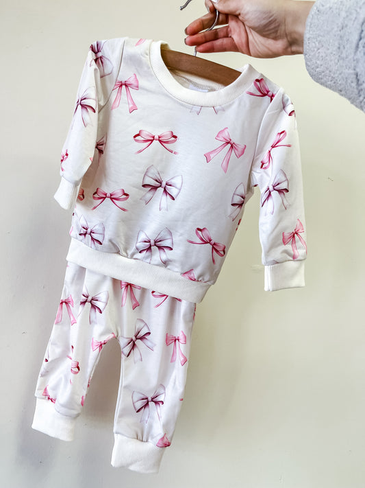 A person holds up the Tiny Tot Bundles Vintage Bow lounge Set, a white baby outfit with pink bows, featuring a long-sleeve top and matching pants.