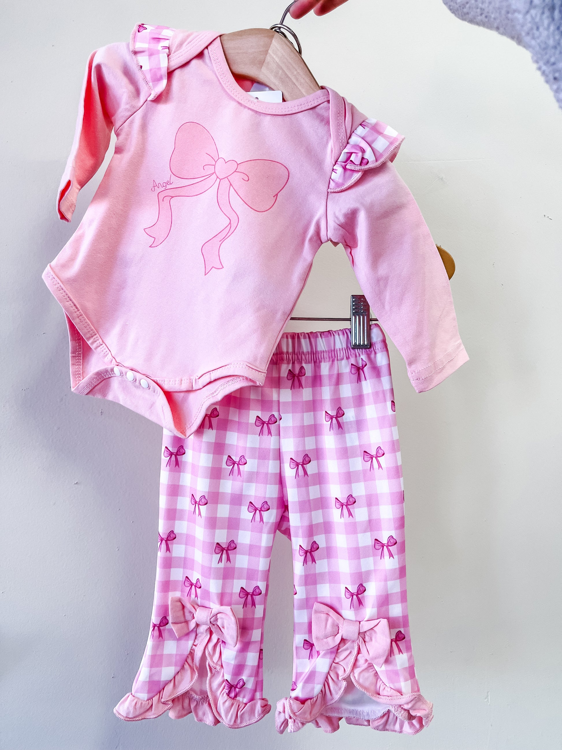 The Angel Bow Blush Set by Tiny Tot Bundles features a pink long-sleeve onesie with a bow and ruffled shoulders, paired with pink-and-white checkered pants adorned with bows and ruffles at the cuffs, displayed on a hanger.